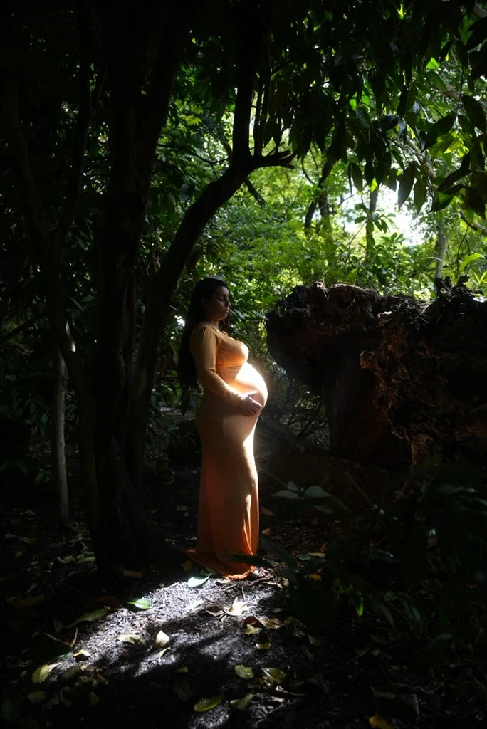 Pregnant woman in a yellow dress standing in a shaded forest with sunlight filtering through trees.