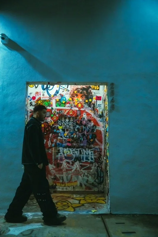 A man walking past a graffiti-covered door with a colorful mural of a woman’s face, illuminated by a blue light in an indoor space.