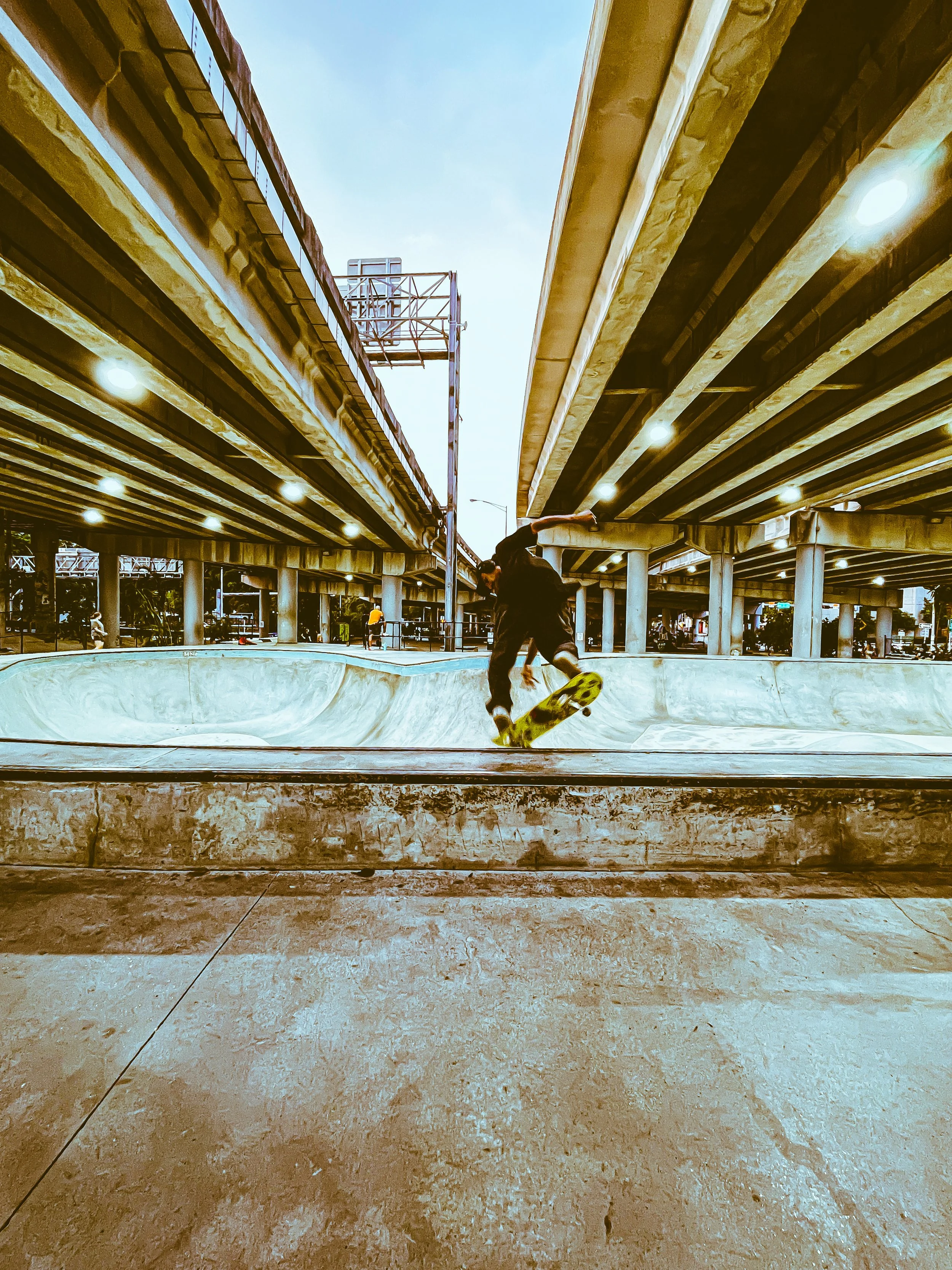 A skateboarder performing a trick in a concrete skate park under an overpass, with bridges and streetlights overhead.