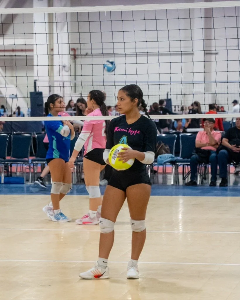 A volleyball player standing on an indoor court holding a volleyball, with other players and spectators in the background.