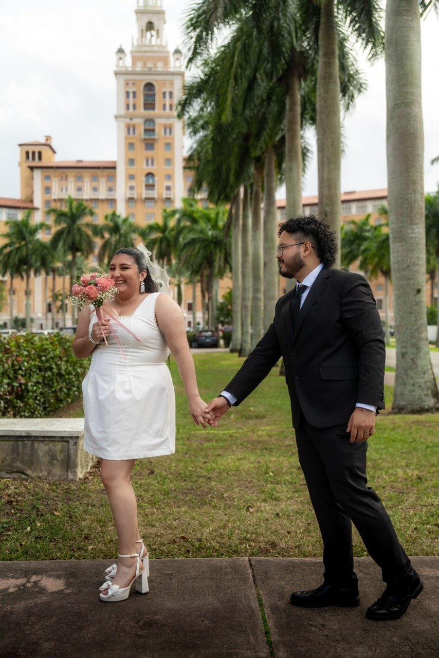 A smiling woman in a white dress and high heels holding a bouquet of pink flowers, being handed by a man in a black suit. They are outdoors with palm trees and a tall hotel building in the background.
