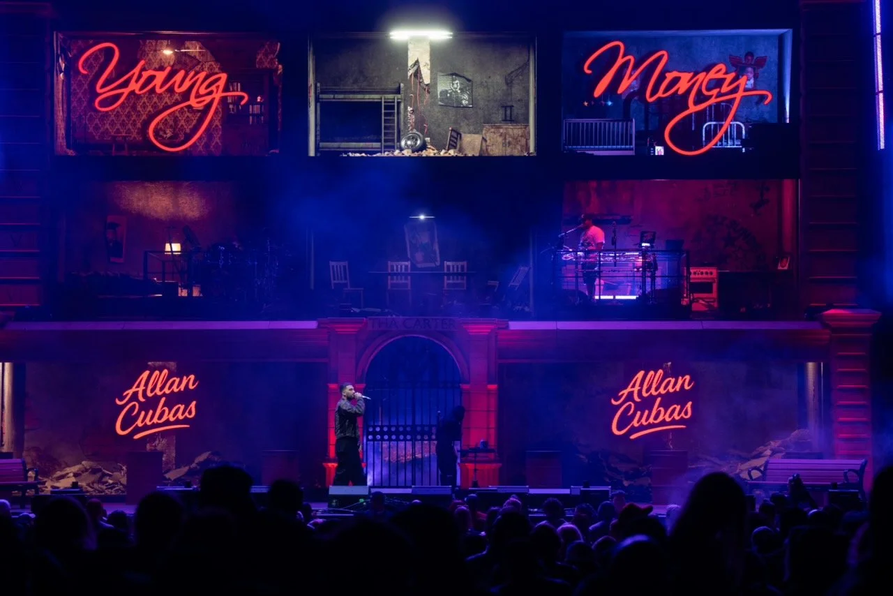 A concert stage with neon signs reading 'Young Money' and 'Allan Cubas', multiple acting areas resembling a house setup with furniture and props, a performer with a microphone on stage, and an audience in the foreground.