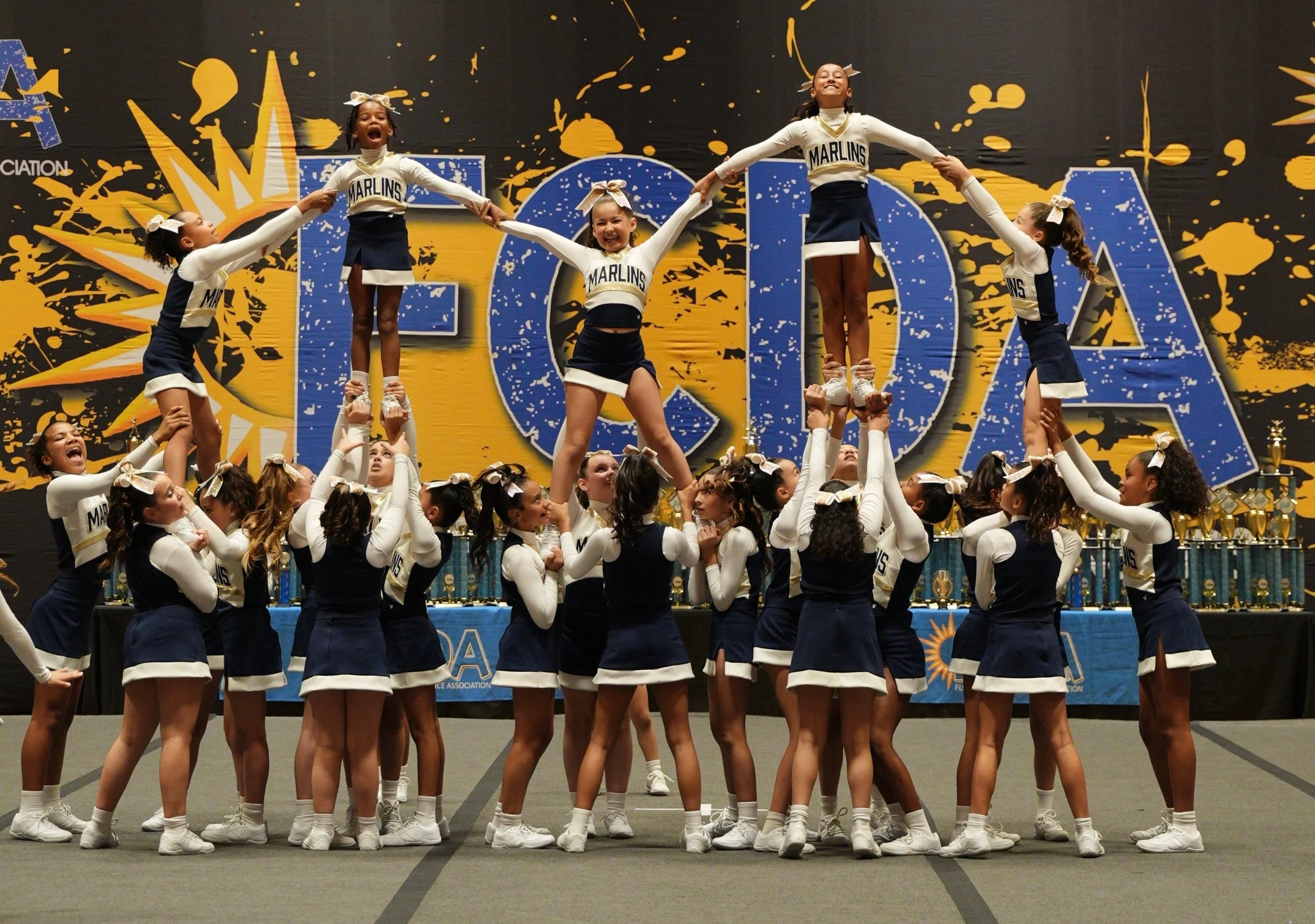 Group of young cheerleaders performing a routine on stage, with some forming human pyramids, wearing navy and white uniforms with 'MARLINS' written on the front, in front of a blue and yellow background with large letters and decorative splashes.