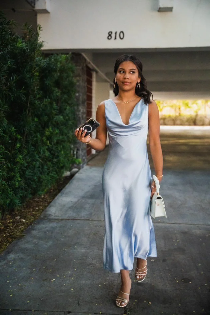 A woman in a light blue satin dress walking outdoors while holding a small white purse and a phone, with greenery on her left and an apartment parking area behind her.