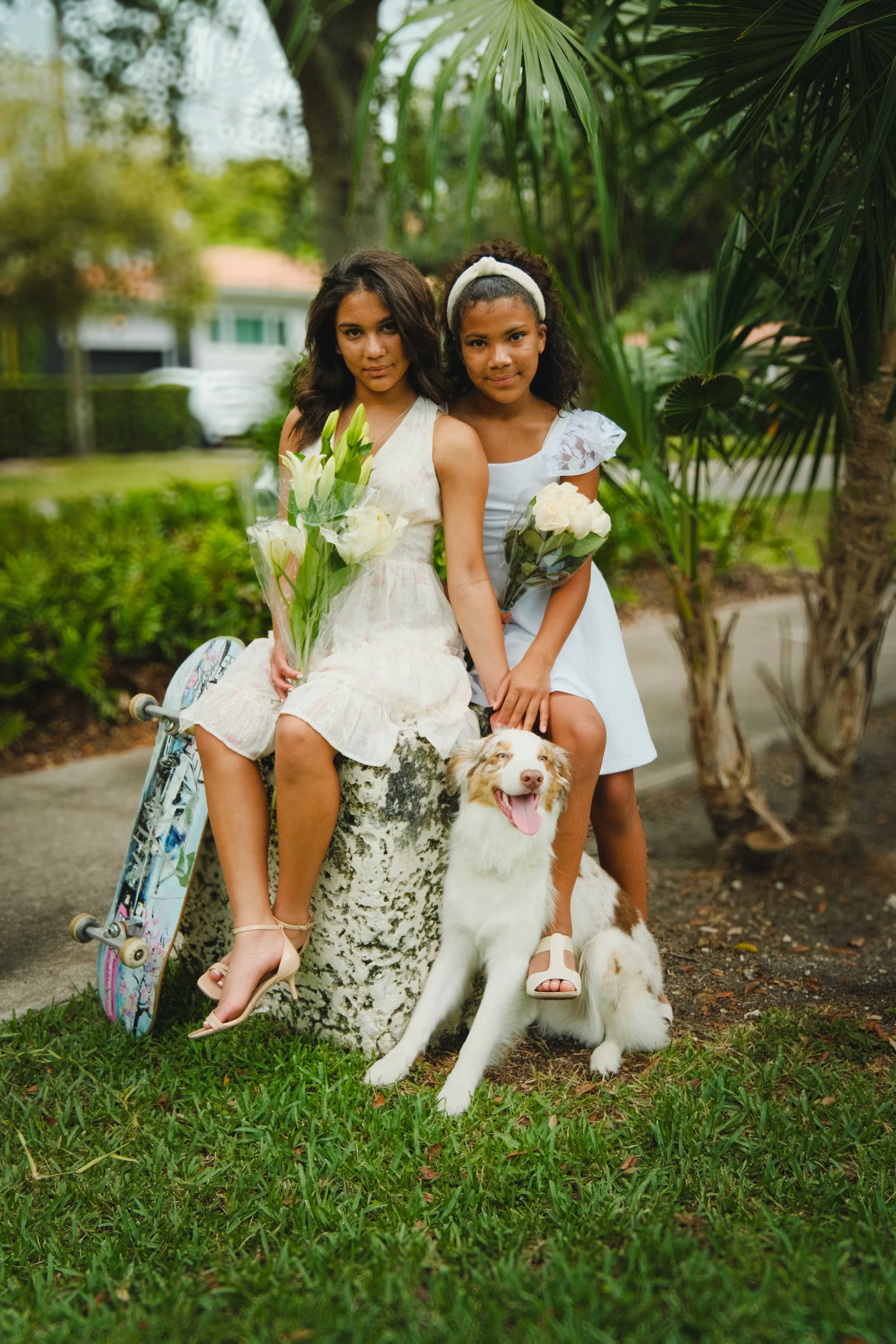 Two young girls sitting on a tree stump in a park, holding bouquets of flowers, with a dog sitting in front of them. One girl is holding a skateboard. There are palm trees and greenery in the background.