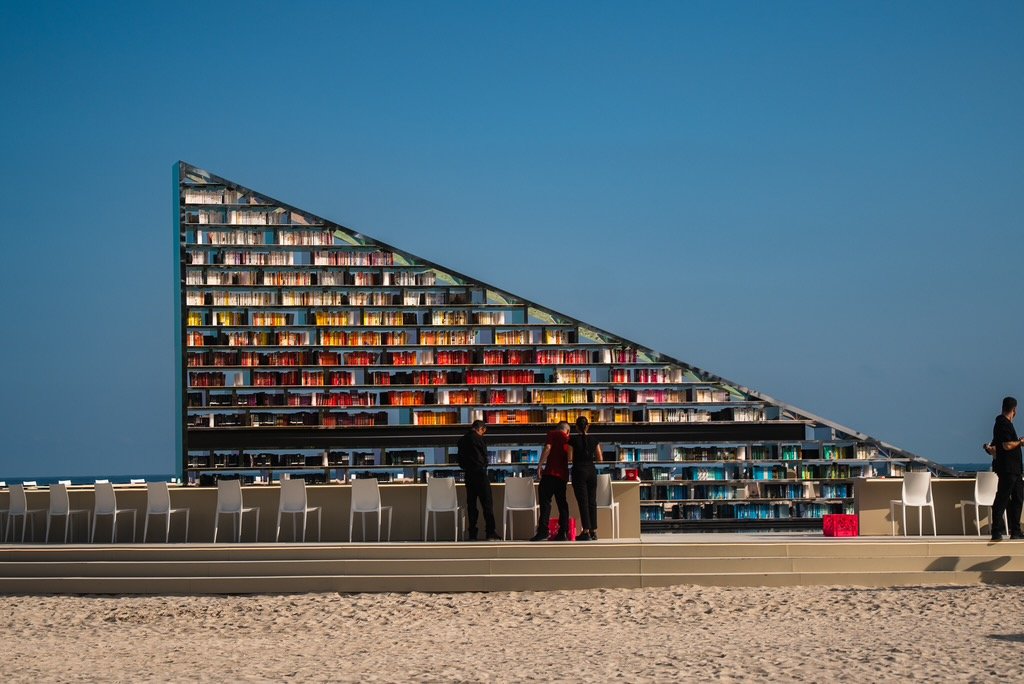 A large, triangle-shaped bookshelf filled with colorful books, located outdoors on a beachfront terrace with a sandy surface, several white chairs, and a few people browsing the books.