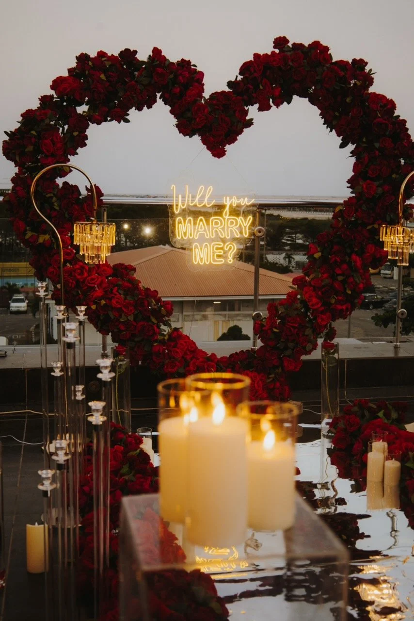 A romantic setting with a large heart-shaped floral arrangement made of red roses, illuminated by candles and chandeliers, with a neon sign reading 'Will you MARRY ME?' in the background.