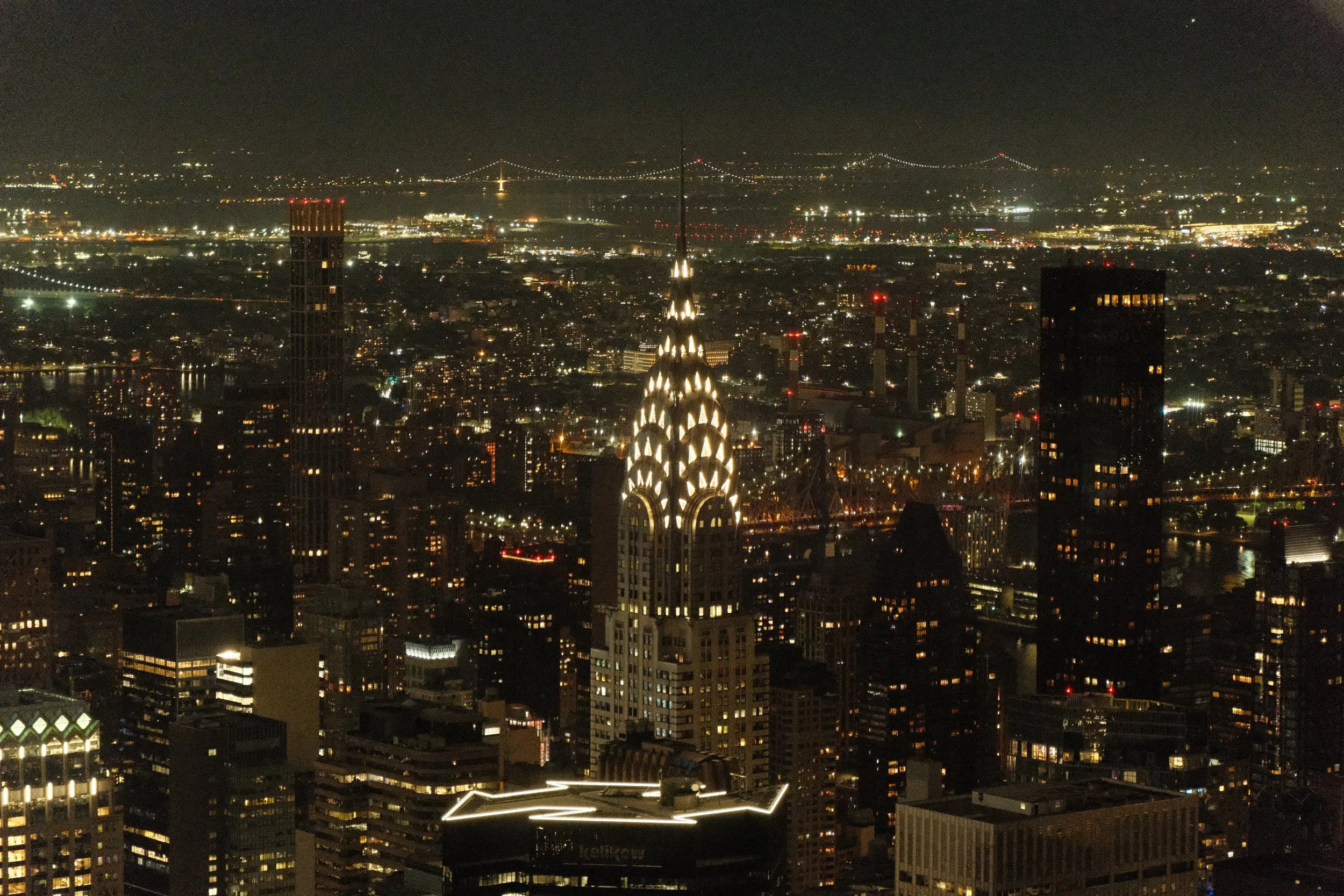 Nighttime cityscape view featuring illuminated skyscrapers and the Chrysler Building in New York City, with a bridge visible in the distance.