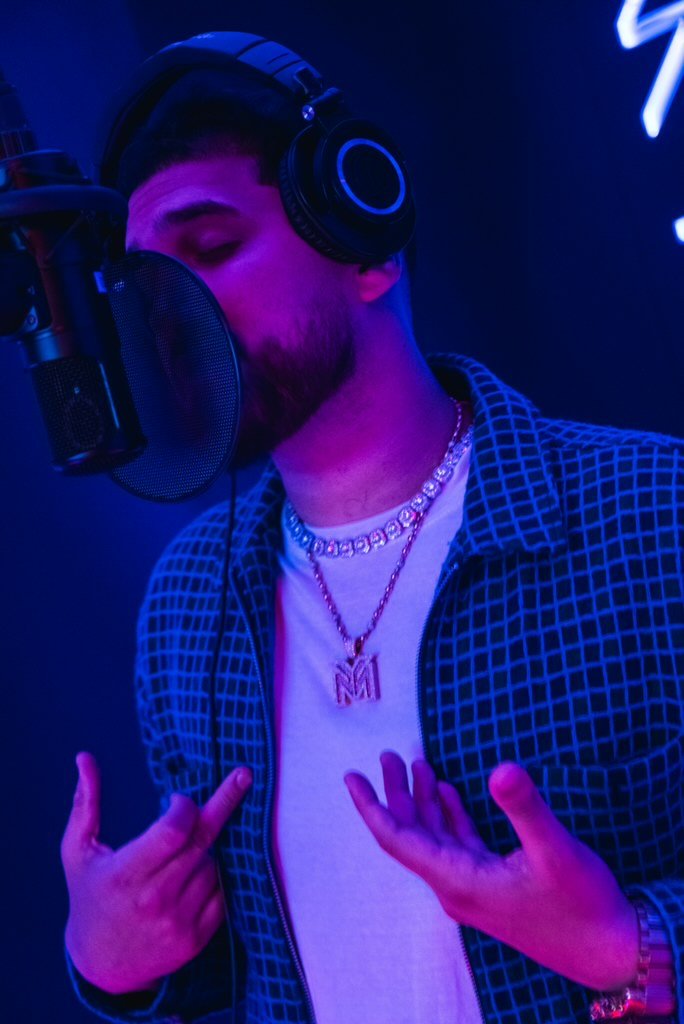 A man with a beard and dark hair in a recording studio, singing into a microphone with headphones, wearing diamond necklaces and a checkered shirt, making a gesture with his hands.
