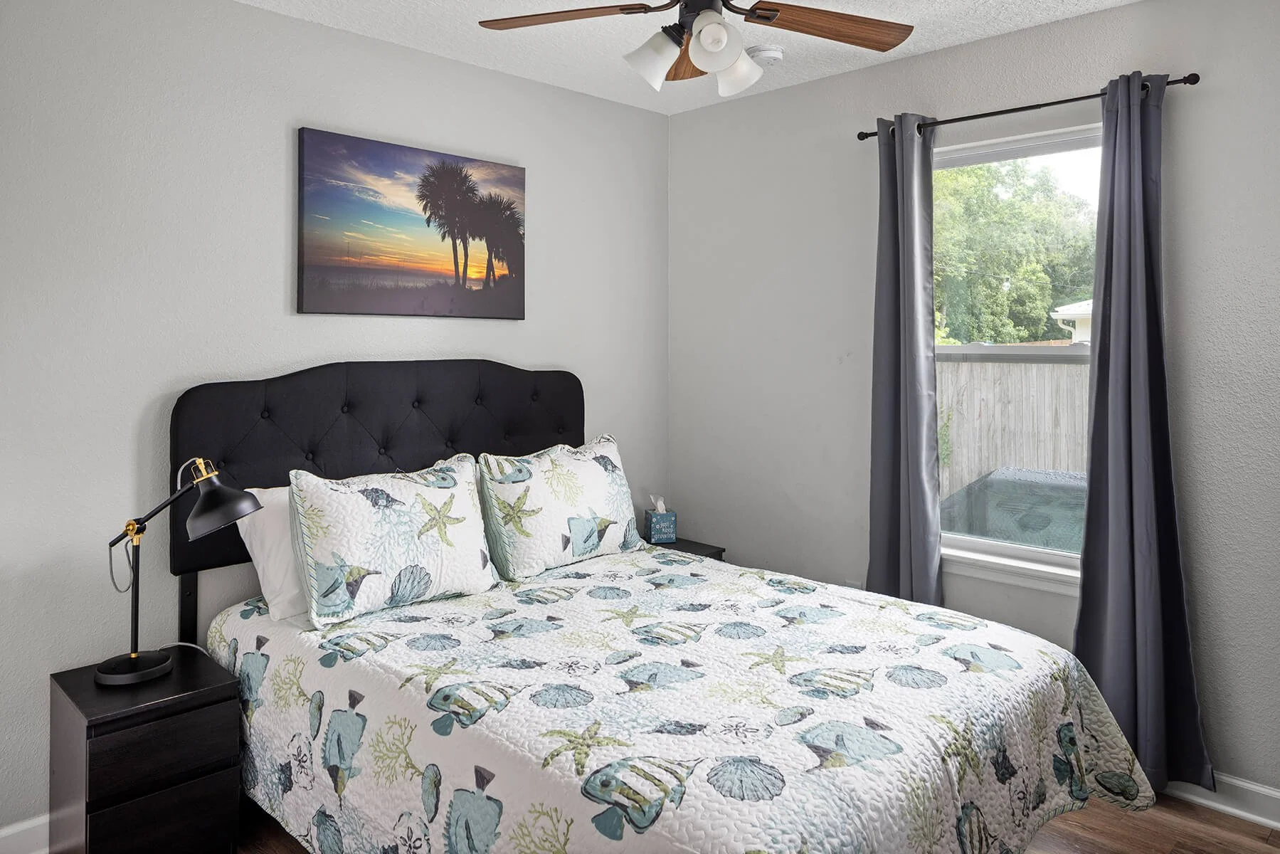 A bedroom with a bed that has a black tufted headboard, decorated with bedding and pillows featuring seashells and starfish patterns. There is a black nightstand with a black desk lamp on the left side of the bed. A window with gray curtains allows natural light to brighten the room, and a ceiling fan with wooden blades and white lights is mounted on the ceiling. A framed sunset picture with palm trees hangs above the bed.