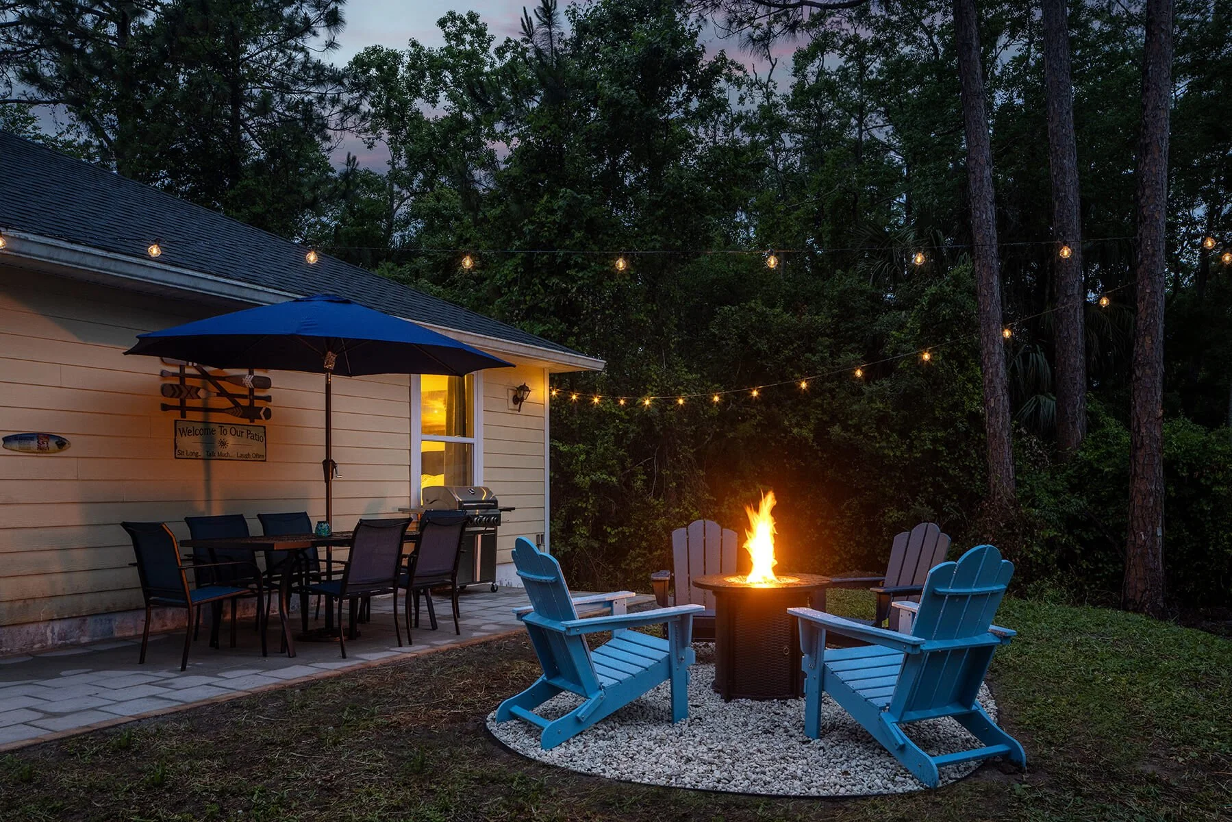 Backyard patio at night with blue Adirondack chairs around a fire pit, string lights hanging above, and a house with beige siding and a barbecue grill.