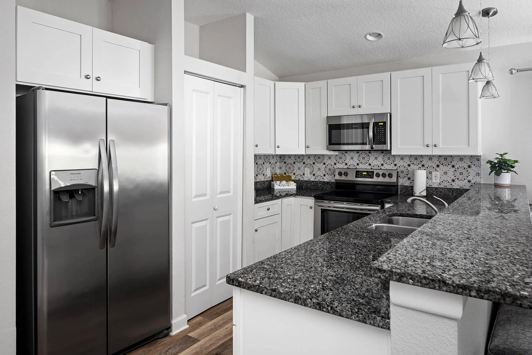 Modern kitchen with stainless steel refrigerator, white cabinets, granite countertops, and patterned tile backsplash. Includes microwave oven, electric stove, sink, and a small potted plant.
