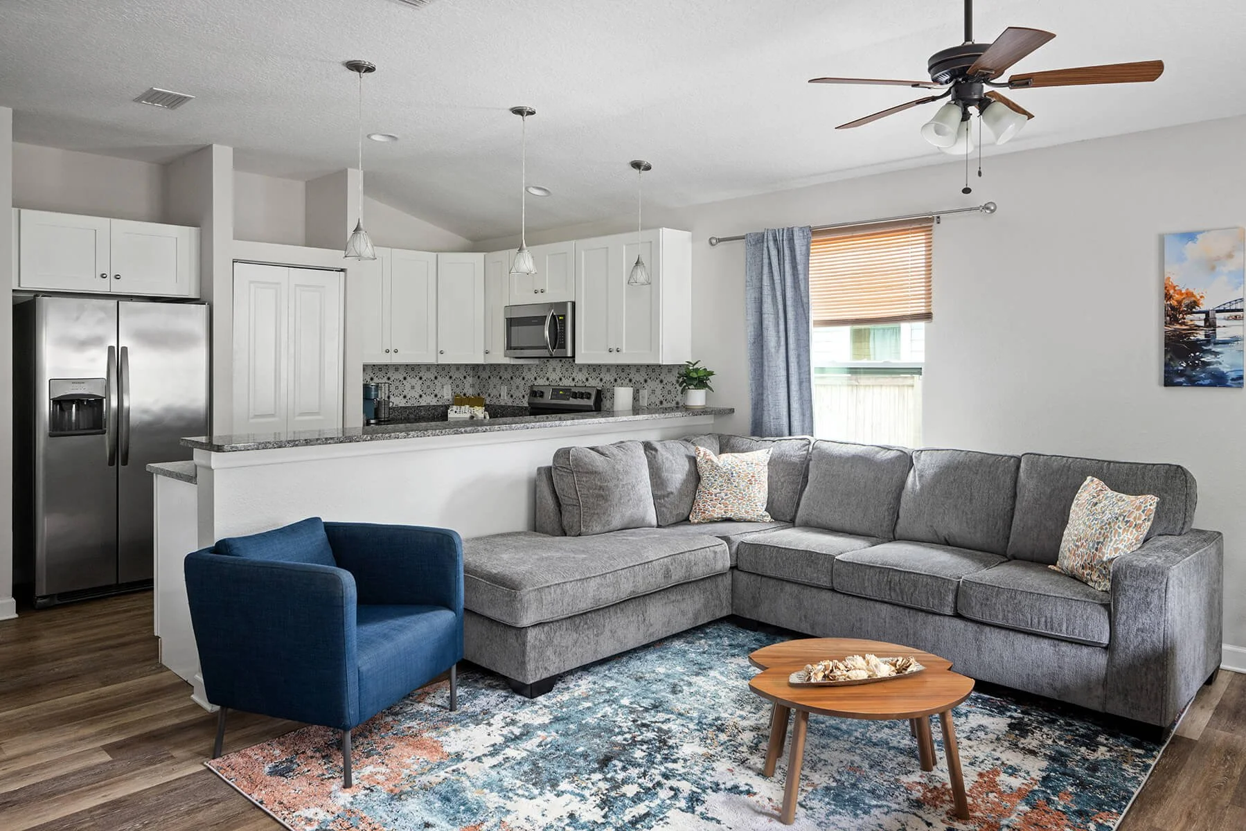 Open-concept living room and kitchen with gray sectional sofa, blue armchair, colorful patterned pillows, wooden coffee table, area rug, white kitchen cabinets, stainless steel refrigerator, microwave, pendant lights, ceiling fan, window with blinds and curtains, and a painting on the wall.