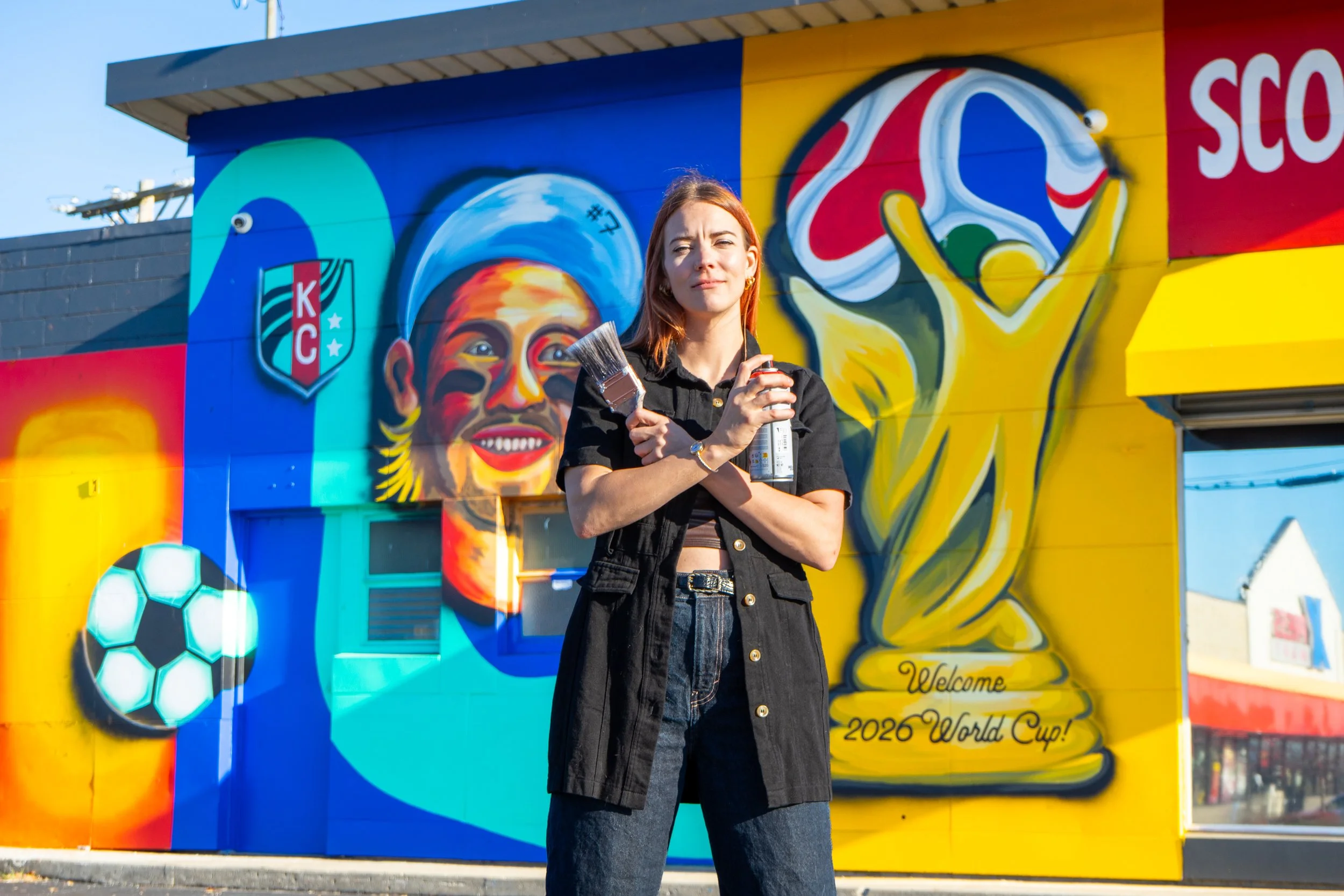 Kansas City muralist and painter, Krysia Badass, standing in front of an outdoor mural she painted for a local sports bar while holding a paint brush and spray paint.