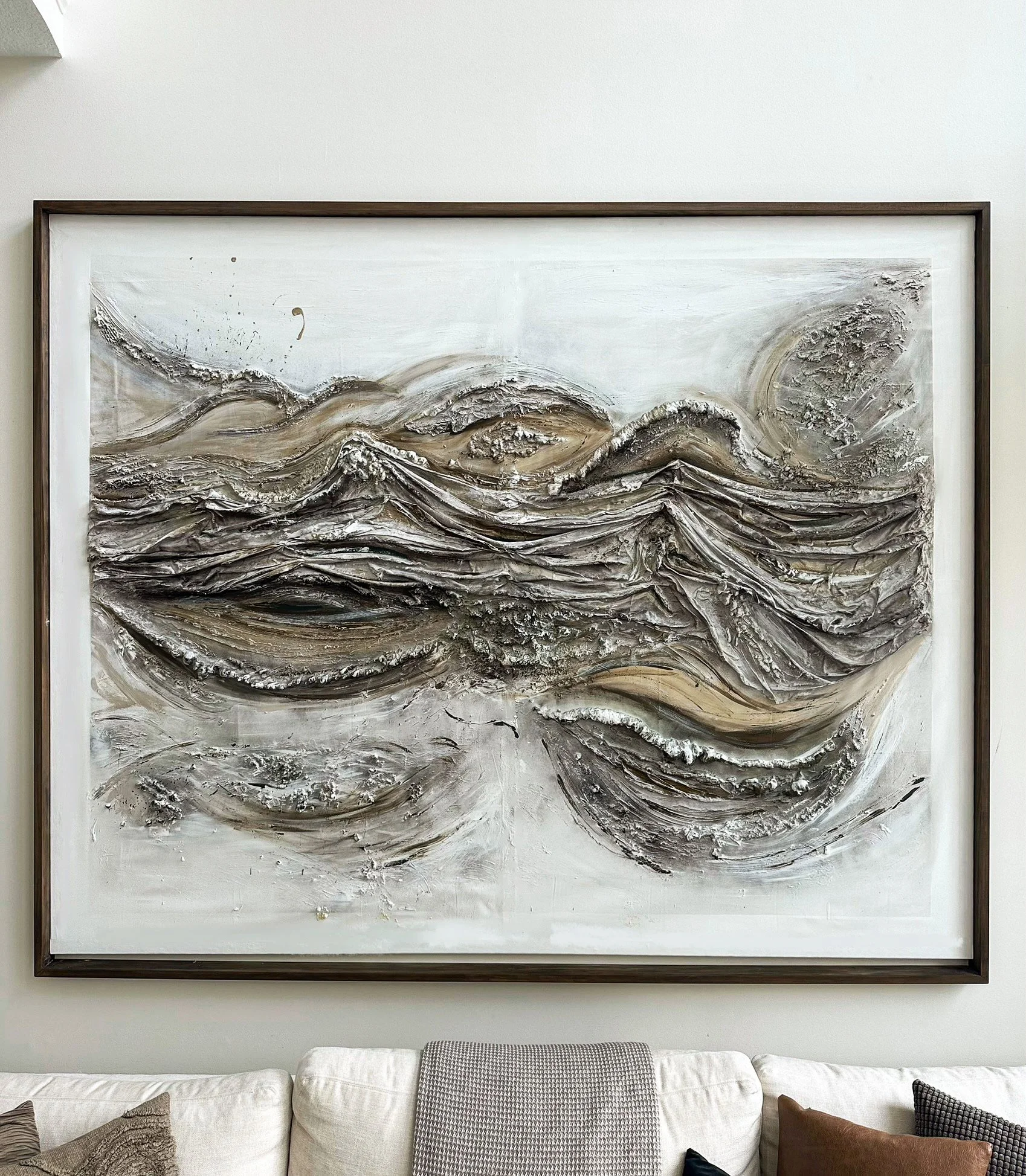 Modern abstract painting with textured, swirling layers in shades of white, gray, and beige, framed and hung on a white wall above a cream-colored sofa with assorted pillows.