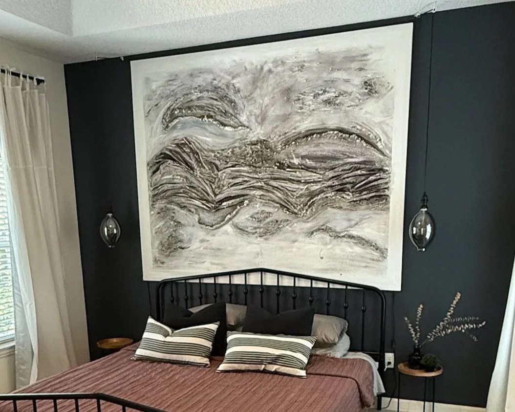 A bedroom with a black metal bed frame, striped and black pillows, and a reddish-brown bedspread. Above the bed is a large abstract black-and-white painting. On either side of the painting are hanging glass pendant lights. To the right, there is a sm