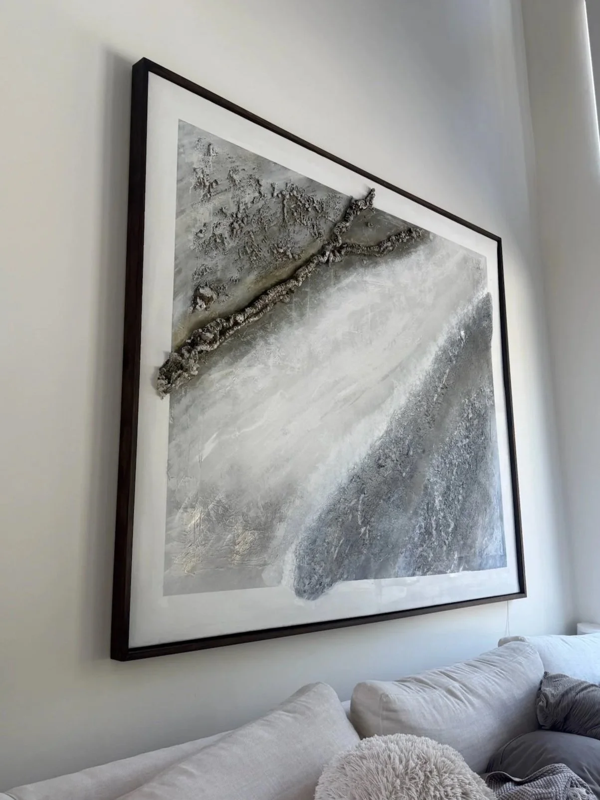 Large abstract wall art featuring textured gray and white patterns and a prominent three-dimensional twisted element, framed in dark wood, hanging above a white sofa.