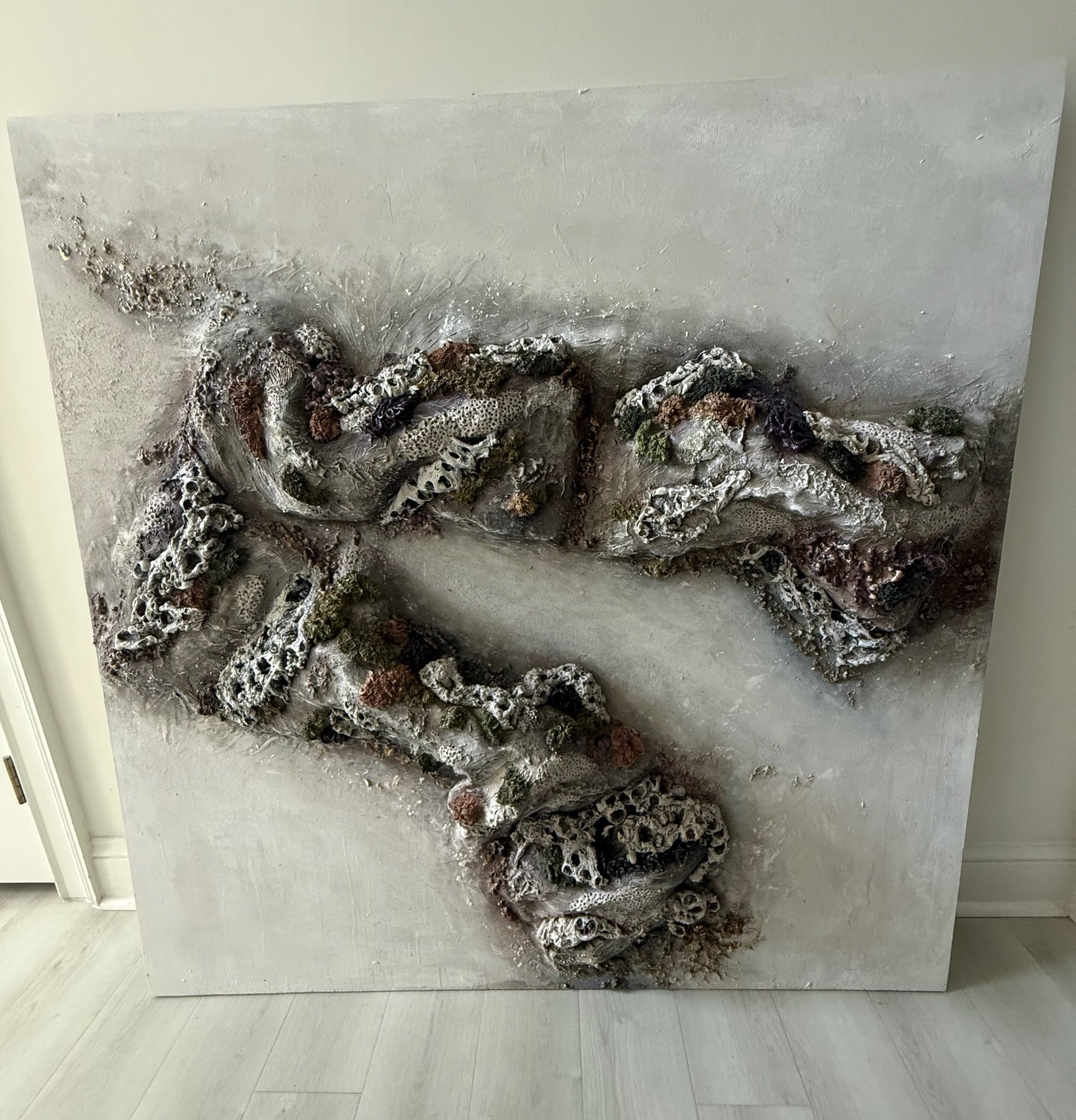 Abstract textured artwork featuring dark, metallic, and earthy colors with organic and coral-like shapes, mounted on a white wall with a light-colored wooden floor below.