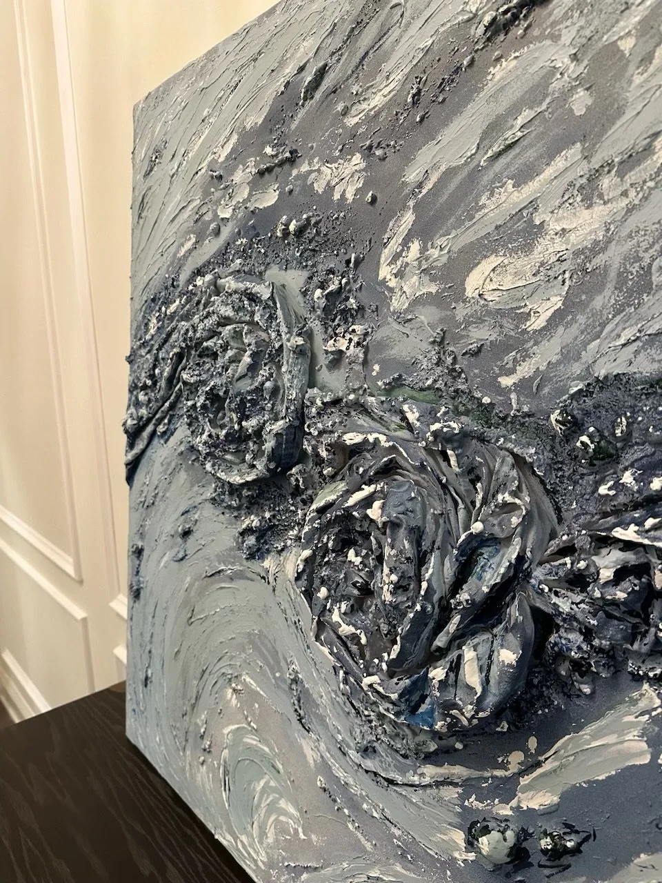 Close-up of a textured, abstract painting with black and gray swirling, three-dimensional rose patterns on a canvas. The painting is leaning against a white wall.