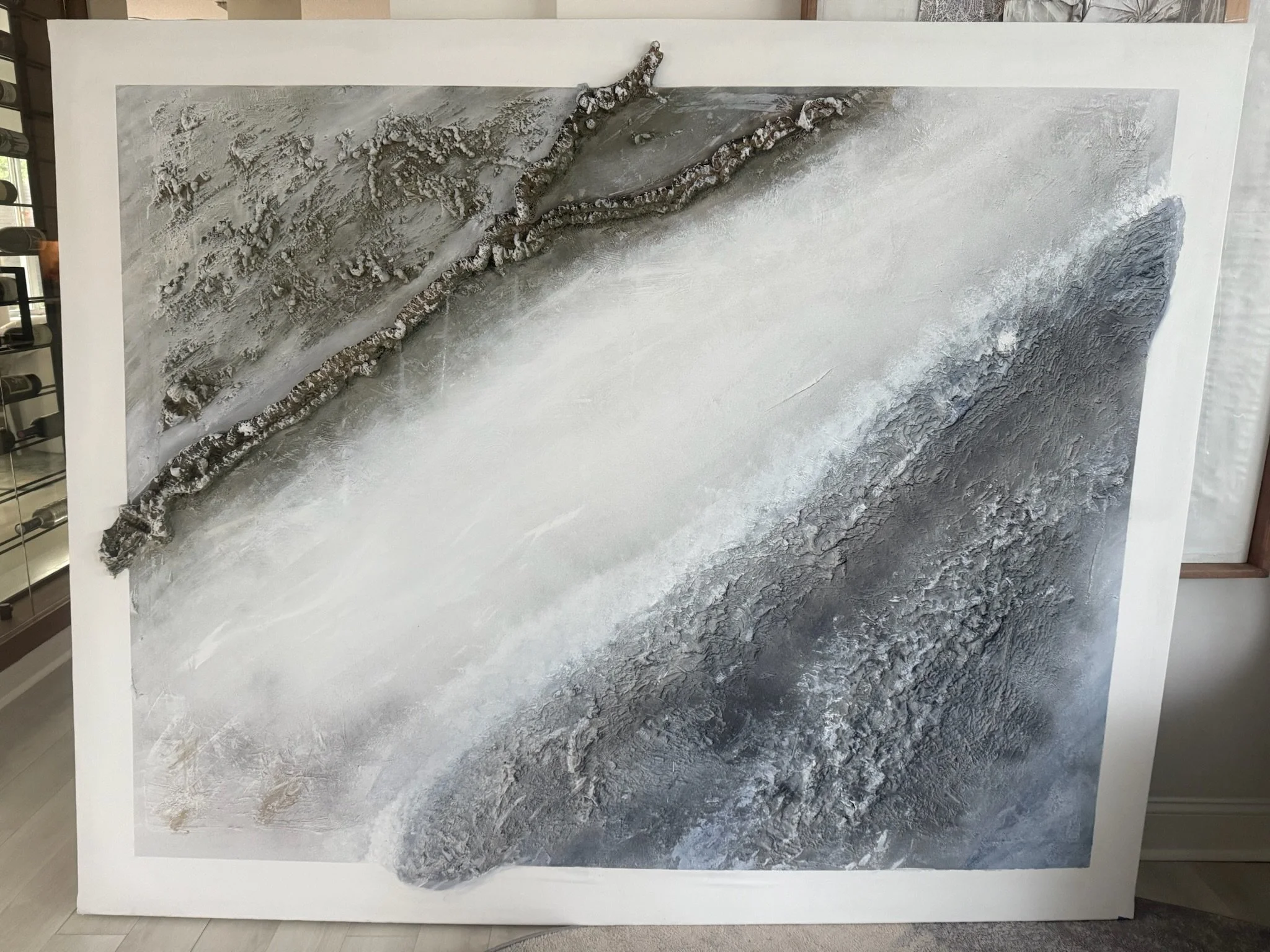 A textured, abstract artwork with shades of gray, white, and black, featuring a diagonal line of rough, bark-like material across the surface.