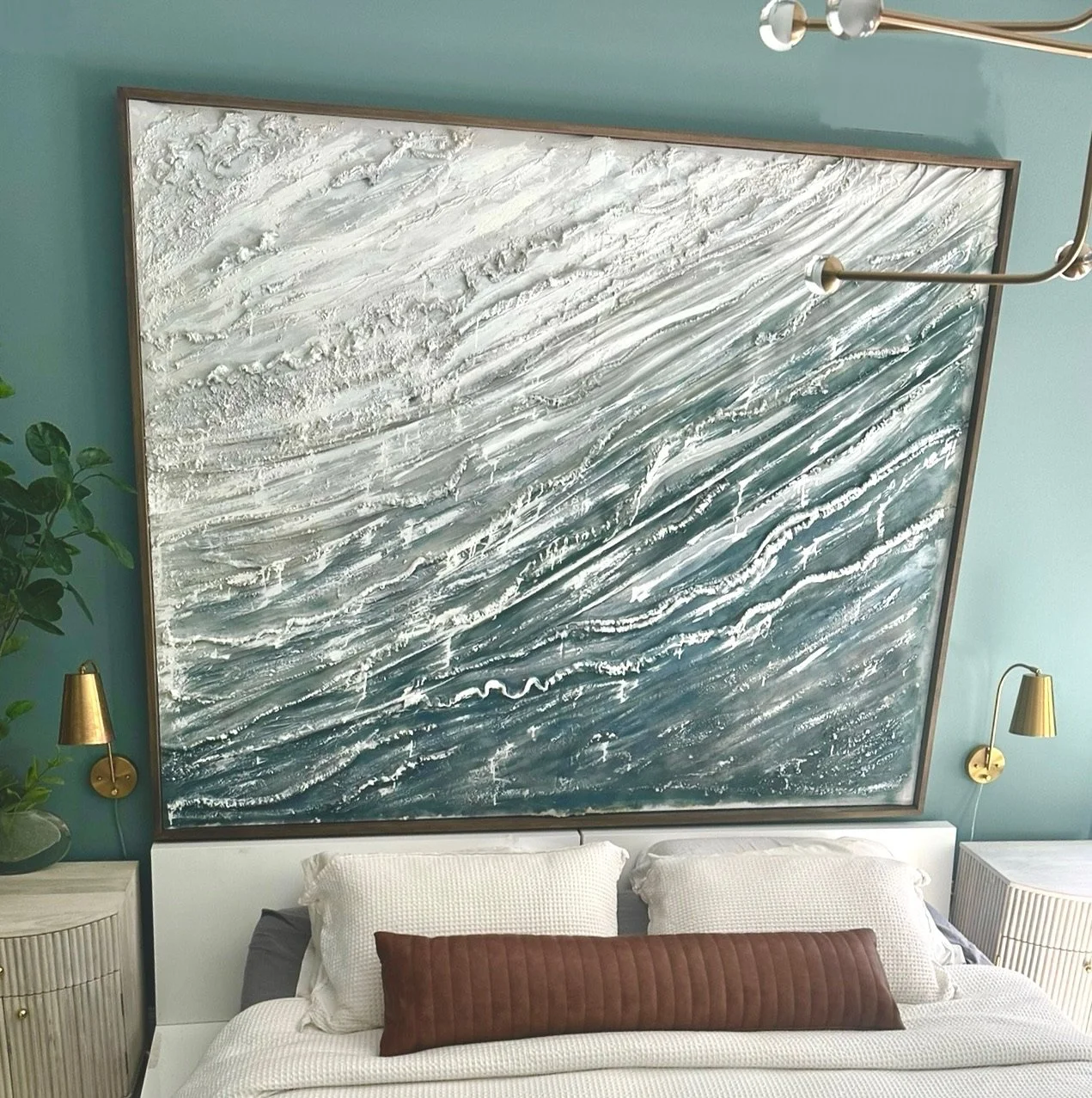 A bedroom with a large, abstract ocean wave artwork above the bed, featuring textures and shades of blue and white, along with wall-mounted gold lamps and a potted plant on the bedside table.
