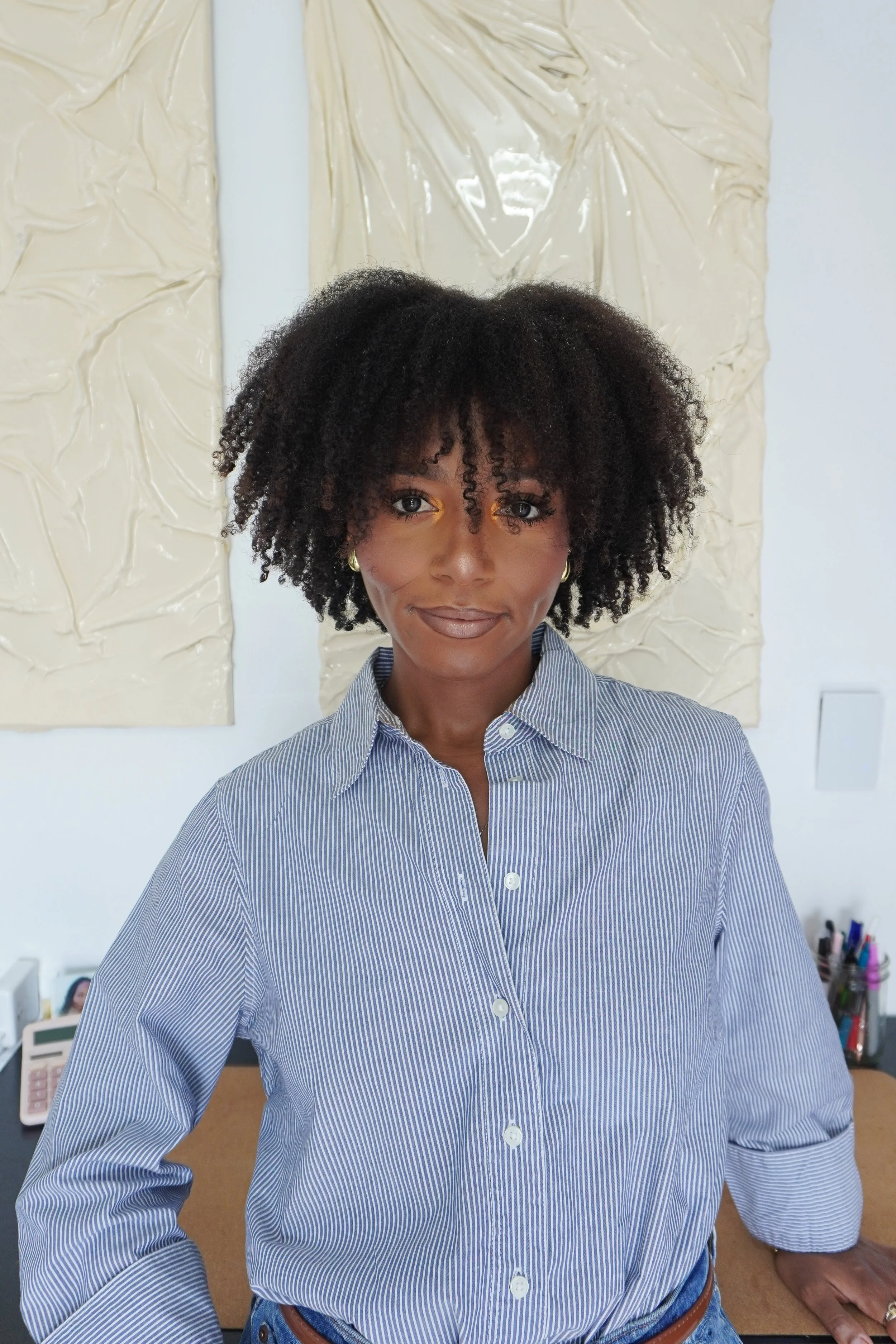 A woman with curly black hair and brown skin wearing a blue and white striped button-up shirt, standing in front of a desk with office supplies and a cream textured artwork behind her.