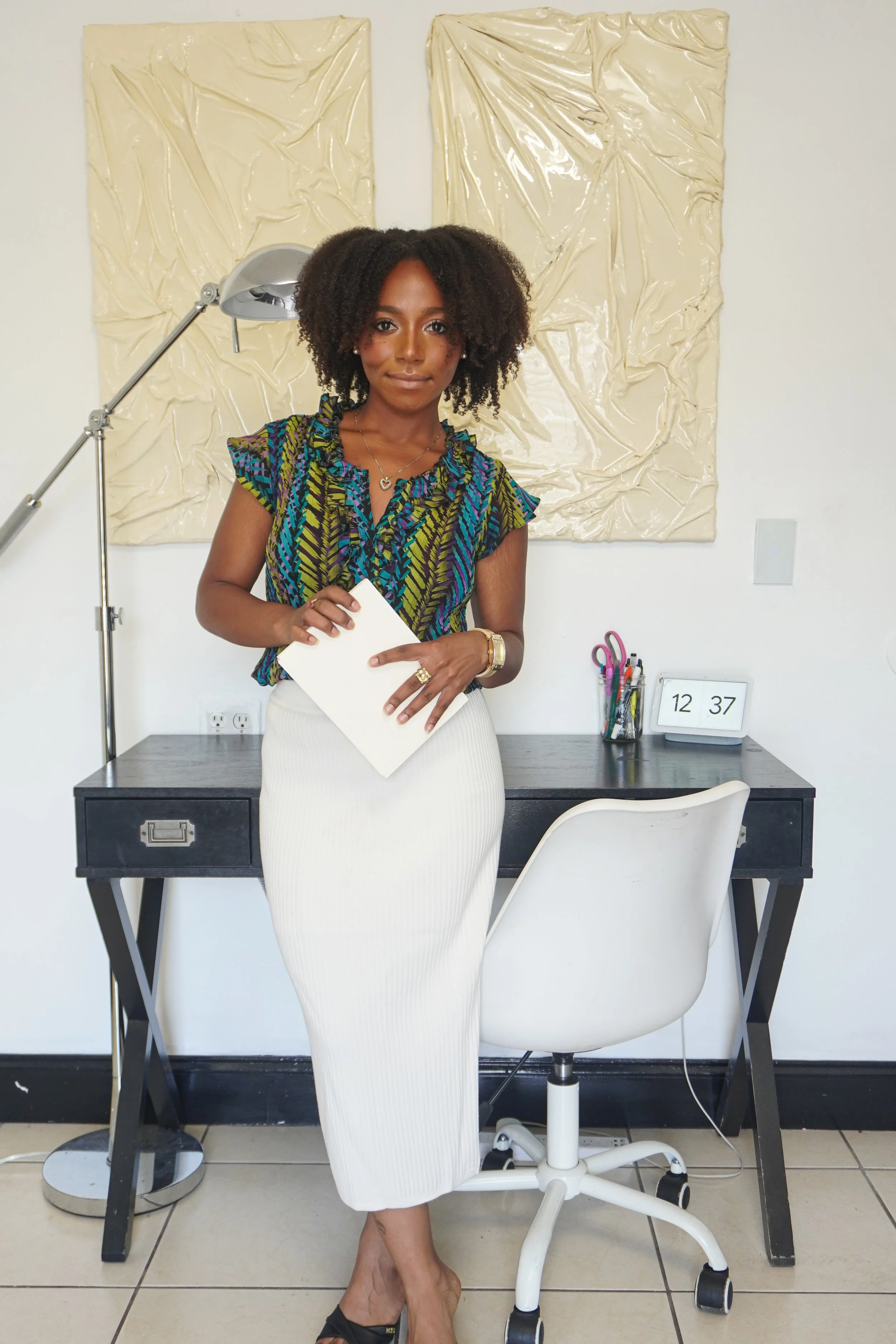 A woman with curly hair wearing a colorful patterned blouse and cream skirt, holding a piece of paper, standing in an office or studio with a desk, chair, and art panels on the wall behind her.