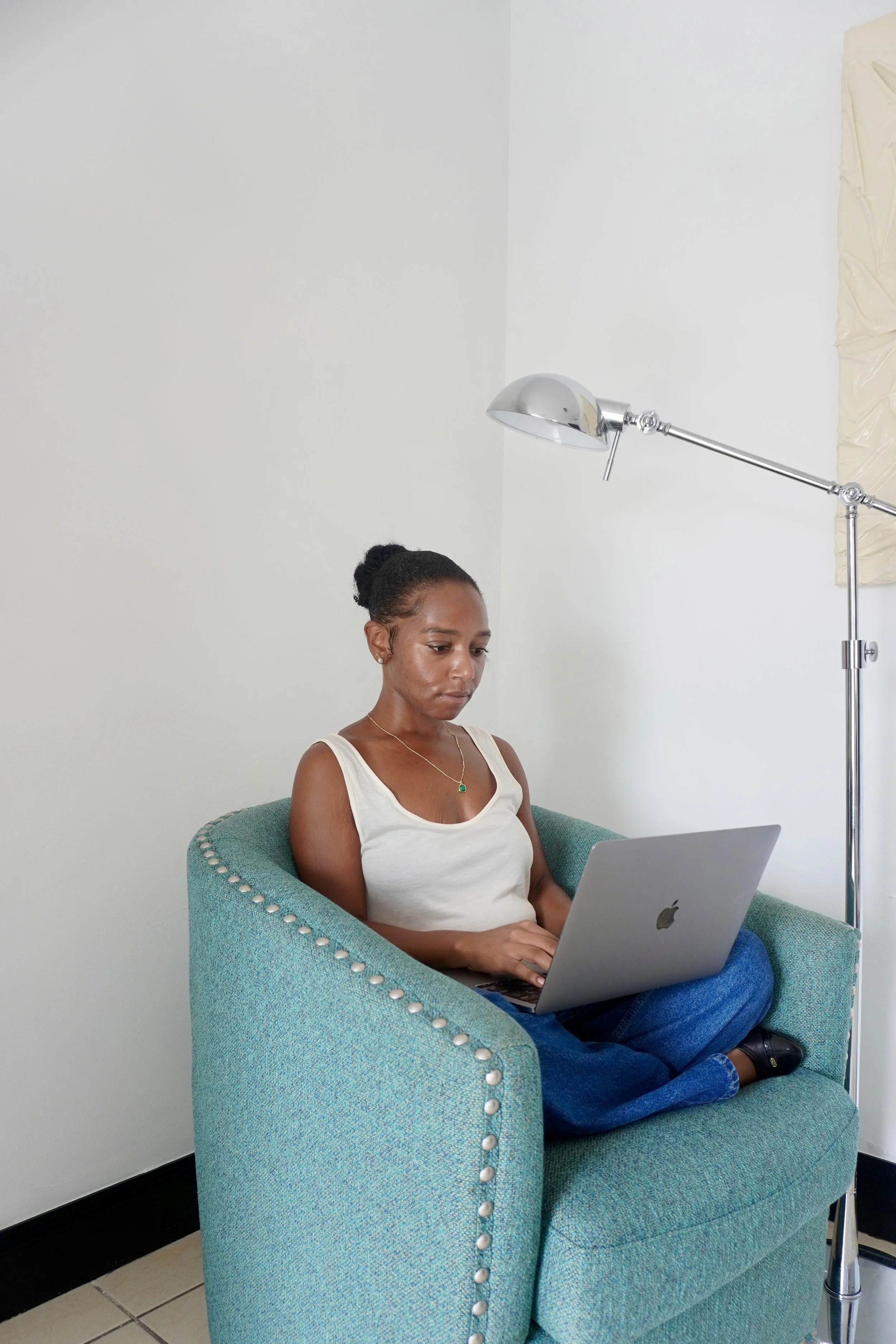 A woman with dark hair tied back is sitting on a teal armchair with white nailhead trim, working on a silver MacBook laptop. She is wearing a white tank top and blue jeans, and appears focused on her work. There is a silver floor lamp next to her and a neutral-colored wall in the background.