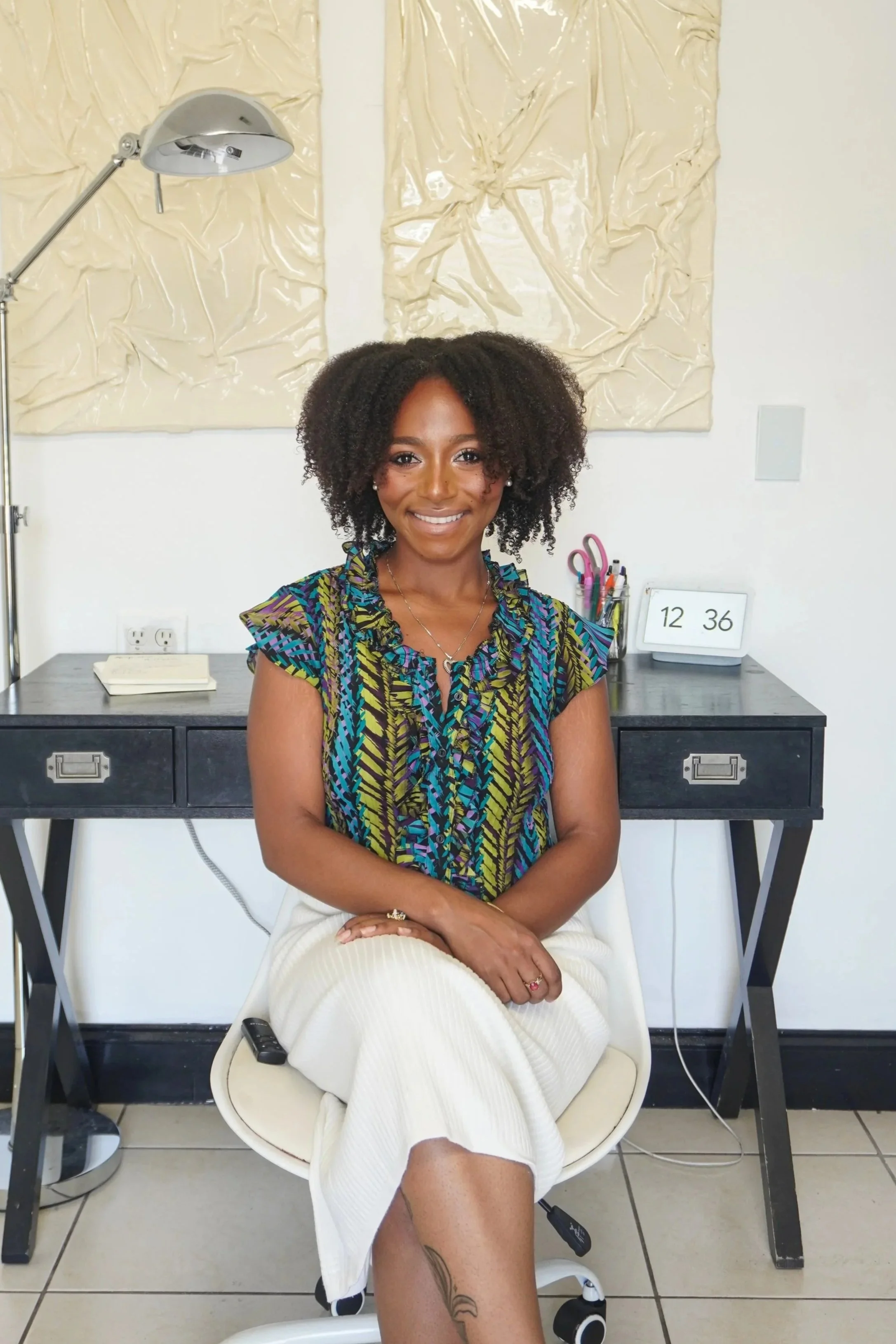 A smiling woman with curly hair sitting on a white chair in an office setting, wearing a colorful patterned blouse and white pants, with her hands resting on her lap. Behind her is a black desk with various office supplies, a clock, and a laptop in a minimalistic room with a beige abstract artwork on the wall.