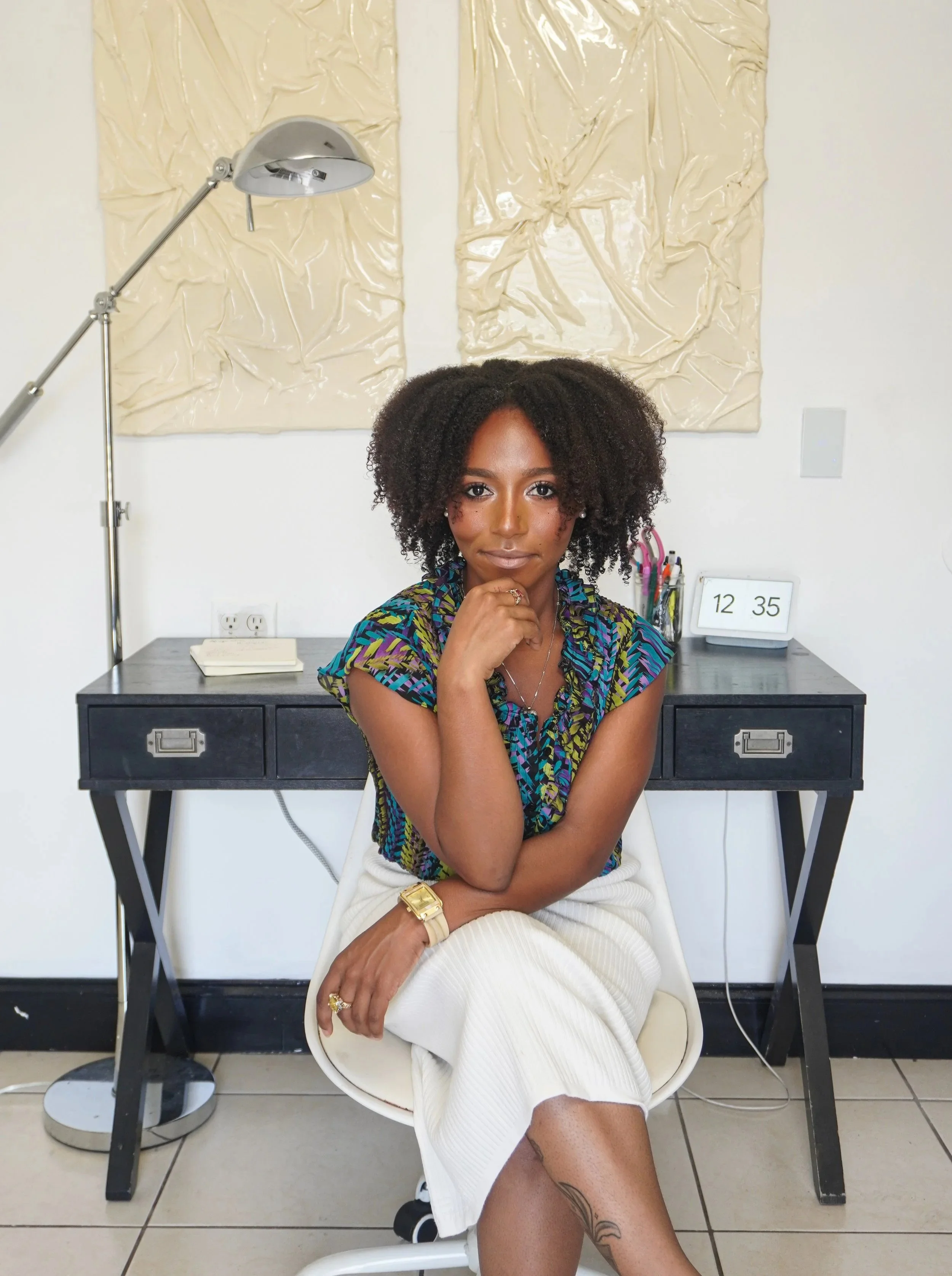 A woman sitting in a white chair in front of a black desk, with her right hand resting on her chin. She has curly hair, a tan complexion, and is wearing a colorful blouse, a gold watch, and rings. There are office supplies on the desk, including notebooks, pens, and a digital clock displaying 12:35. Behind her, a wall with abstract beige artwork and a tall, silver desk lamp are visible.