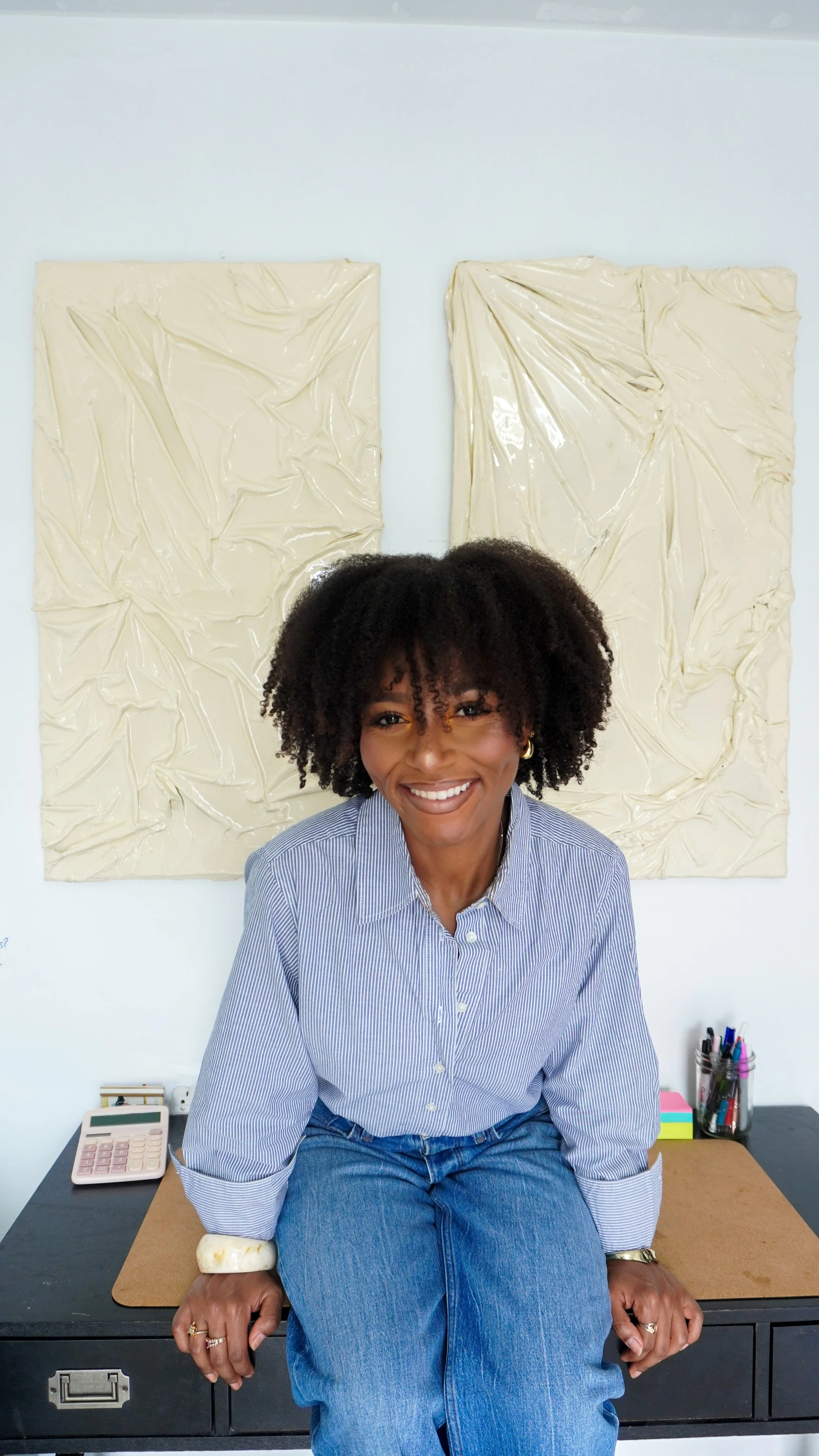 A smiling woman with curly hair and gold earrings sitting on a desk in an office. She is wearing a blue and white striped shirt, jeans, and a white bracelet. Behind her are two abstract cream-colored artworks on the wall. The desk has a calculator, some pens, and other office supplies.