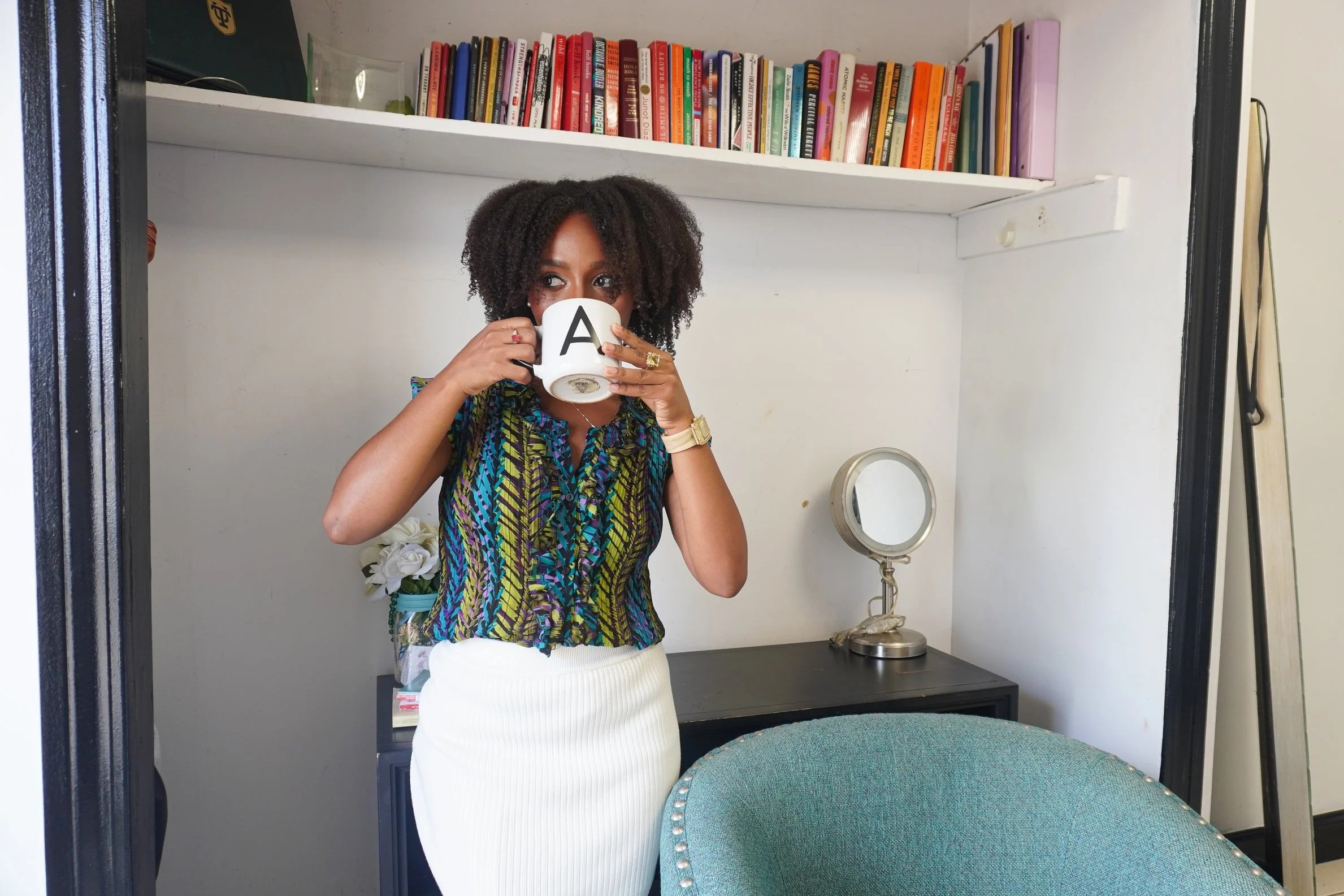 A woman with curly black hair wearing a colorful patterned blouse and white skirt, standing in front of a mirror, holding a white mug with a black letter 'A', inside a room with a bookshelf and a table mirror.