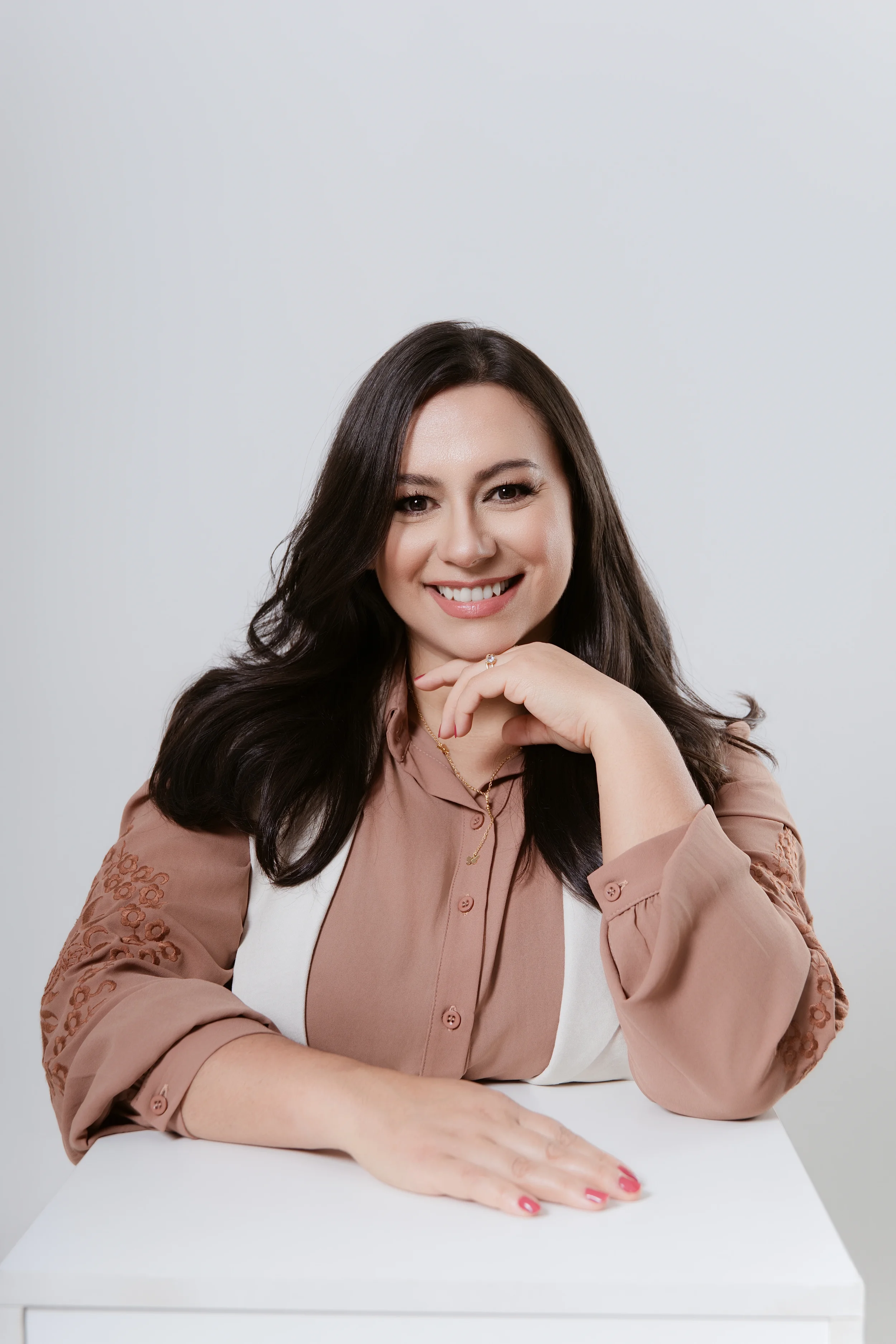 Mulher sorrindo com cabelo castanho, pele clara, usando blusa rosa com detalhes de renda na manga e acessórios dourados, posando com a mão no queixo, diante de fundo branco.