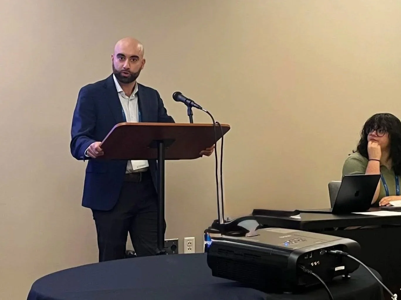 Man with a shaved head and beard wearing a dark suit, white shirt, speaking at a podium with a microphone, while a woman with black hair and glasses sitting at a table with a laptop looks on.