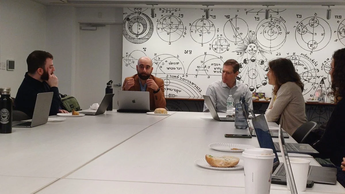 A group of five people sitting at a conference table in a meeting room, engaged in discussion. The background features a wall with scientific diagrams and symbols.