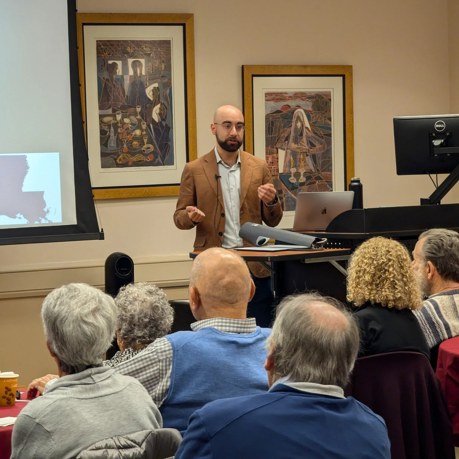 A man in a tan blazer giving a presentation in front of an audience. Behind him are two framed art pieces on the wall. The audience consists of older adults seated around tables, some with beverages, and attentively listening.