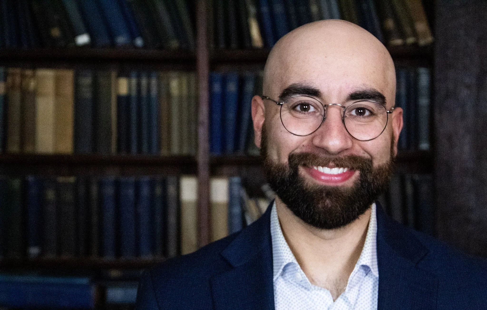 Close-up of a smiling man with glasses and a beard, wearing a suit jacket and shirt, standing in front of a bookshelf filled with books.