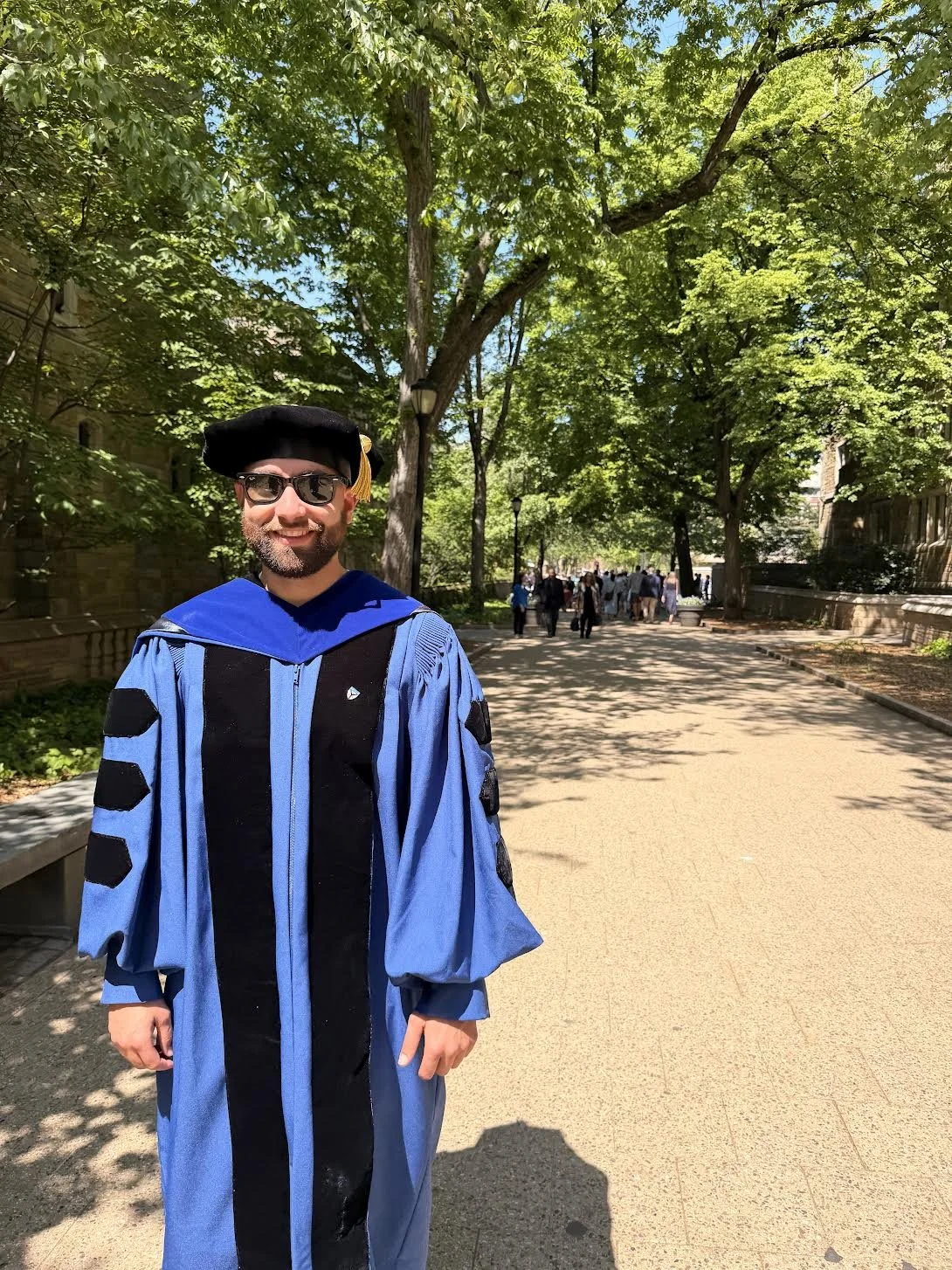 Man in blue graduation gown and cap standing on a sunny tree-lined pathway with other people in the background.