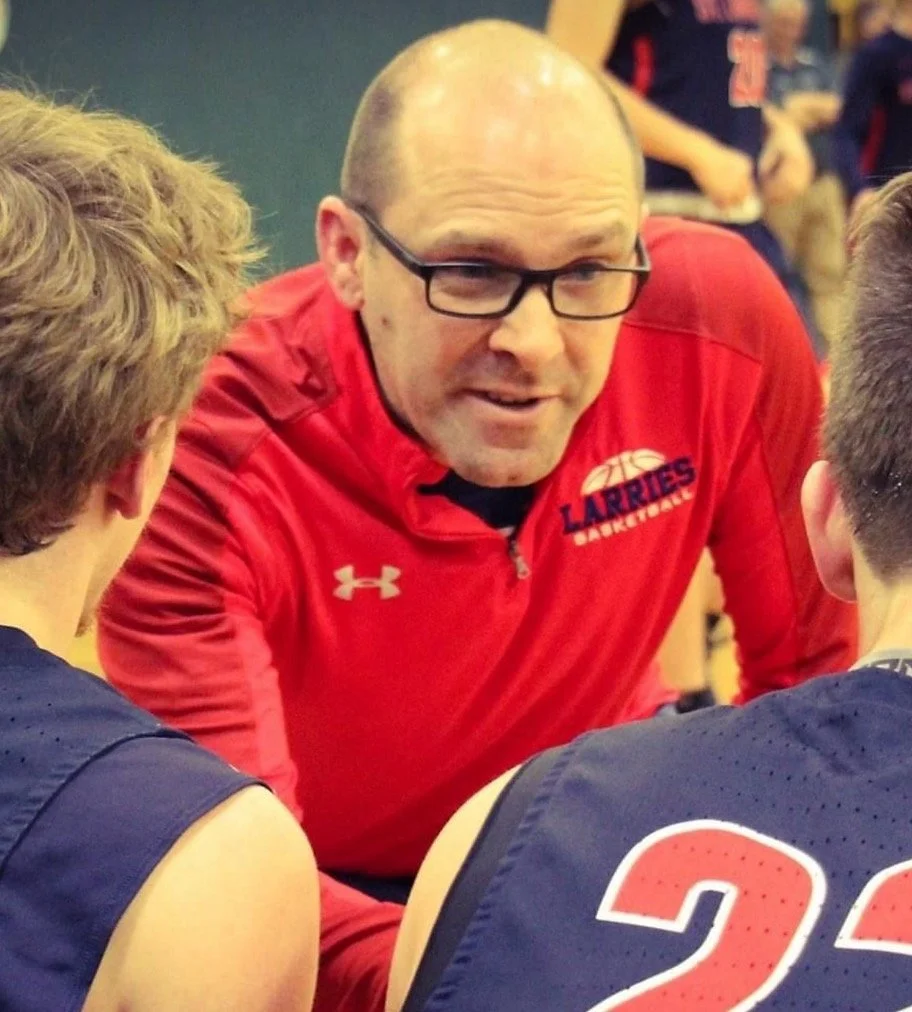 A basketball coach wearing glasses and a red shirt with a basketball logo, speaking to two young basketball players in dark jerseys with red numbers, in a gym setting.