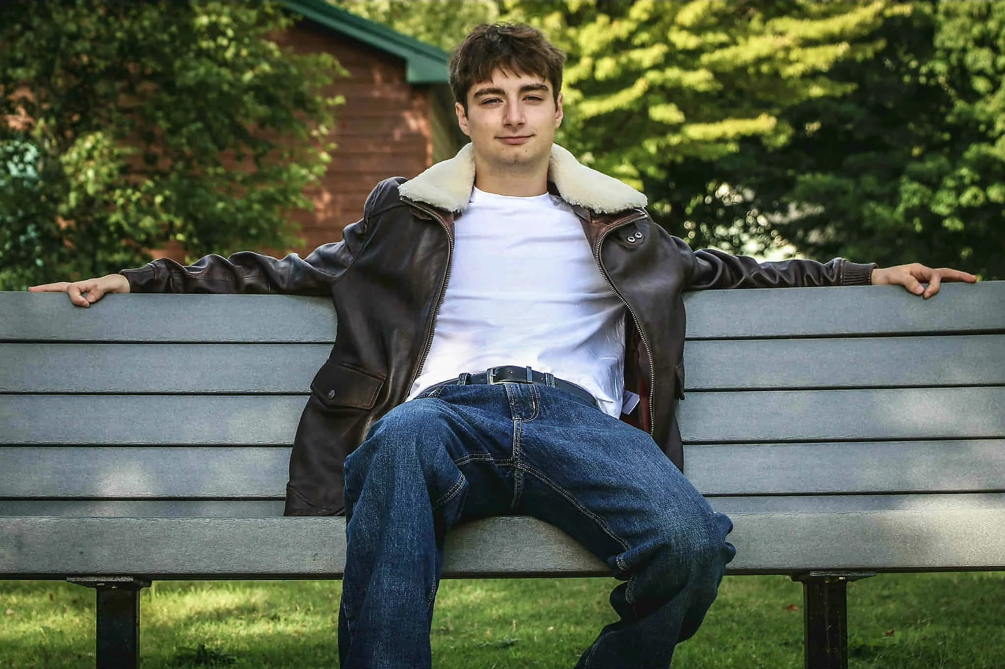A young man sitting on a park bench with arms outstretched, wearing a leather jacket with a shearling collar and blue jeans, in front of green trees and a small wooden structure.
