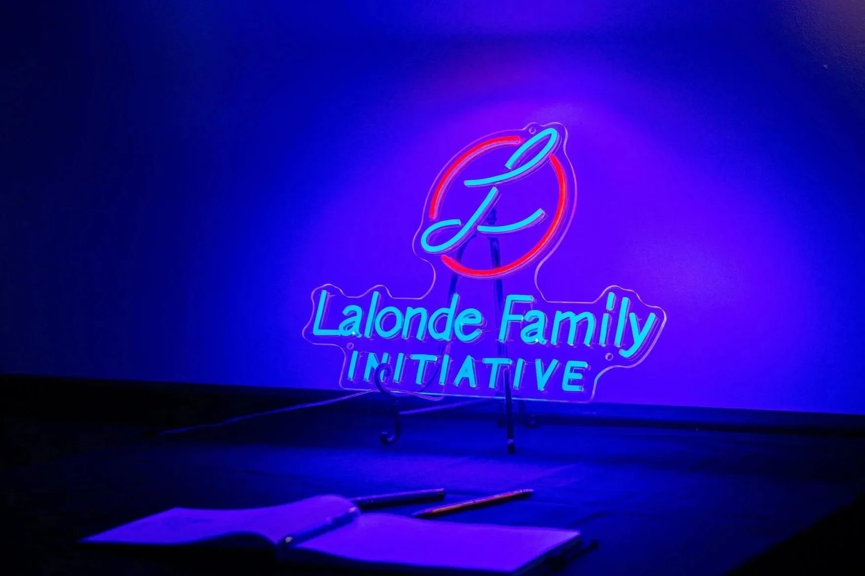 Neon sign reading 'Lalonde Family INITIATIVE' with a circular design and a stylized letter 'L' in the center, glowing in blue and red hues, against a dark background.