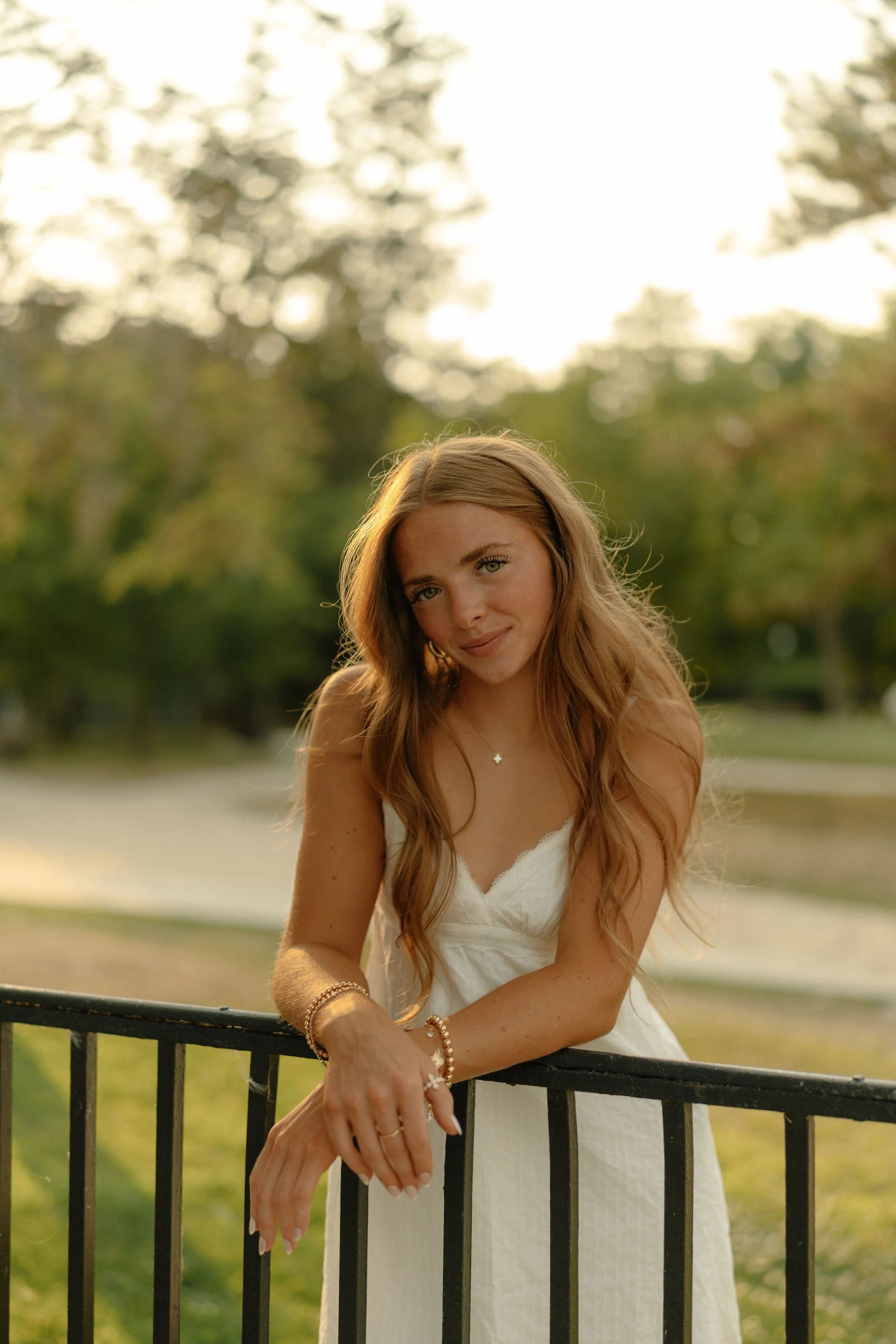 Young woman with long wavy red hair wearing a white dress, standing outdoors near a black metal railing with green trees and bright sky in the background.
