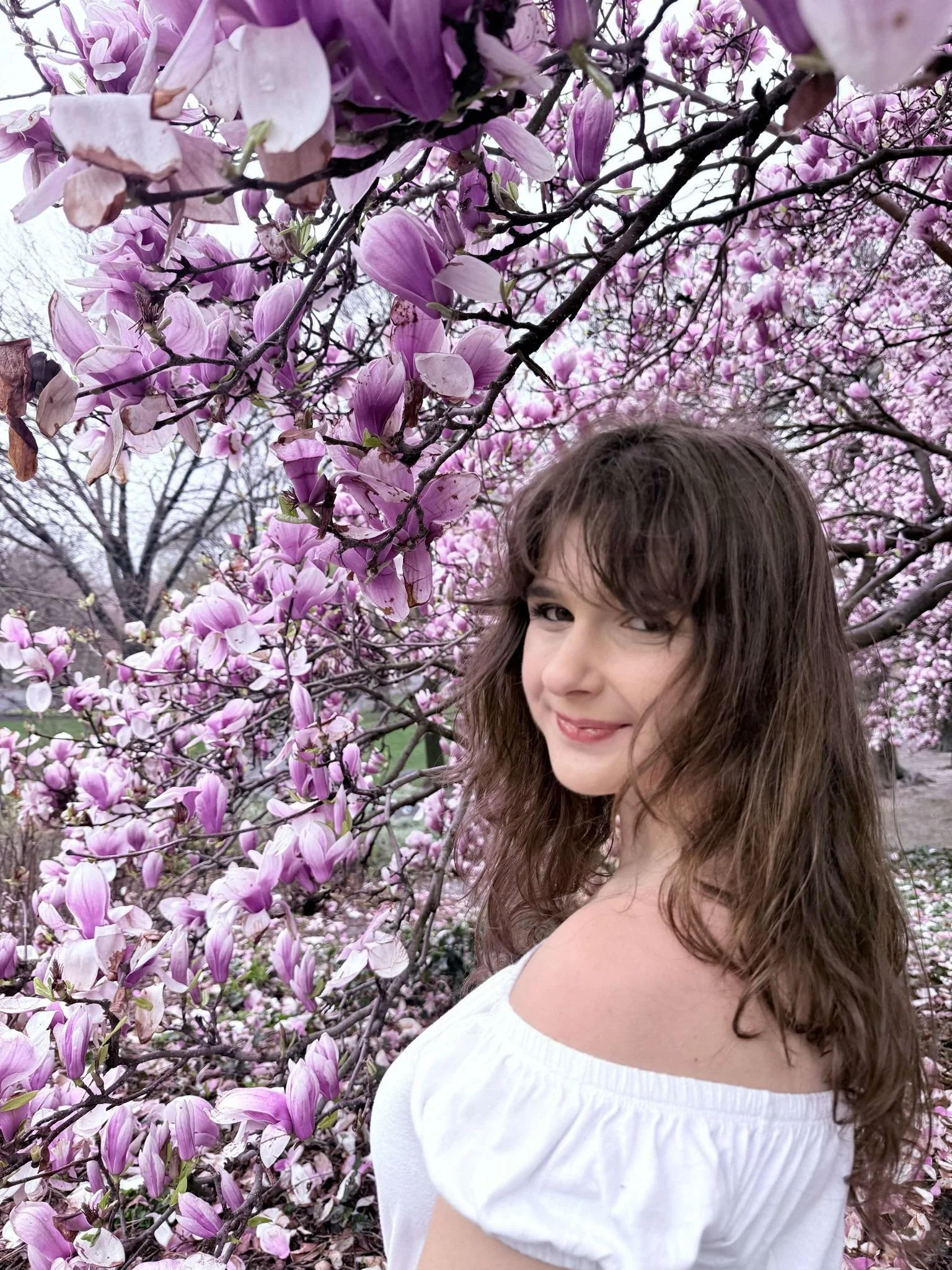 A woman with brown hair and a white off-shoulder top standing in front of pink blooming magnolia trees.