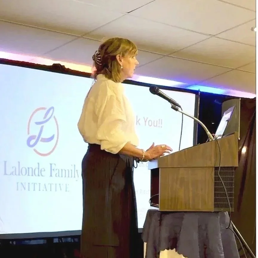 A woman standing at a podium giving a presentation or speech, with a large screen displaying a logo and text that says "Lalonde Family Initiative" and "Thank You!!" in the background.