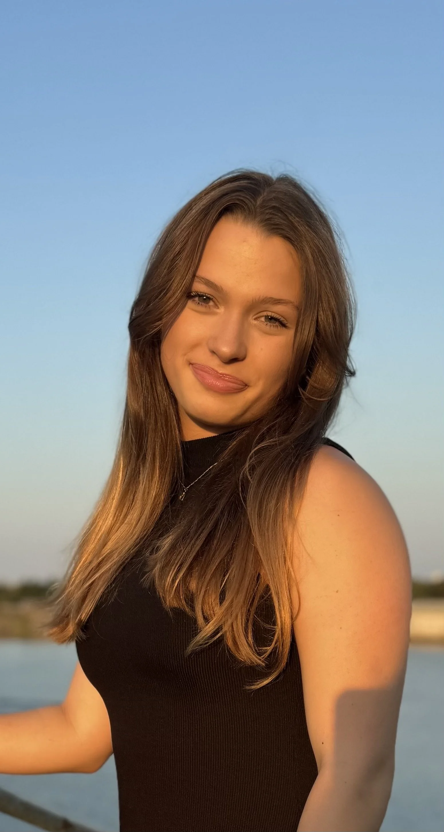 A young woman with long brown hair, smiling, wearing a black top, outdoors near a body of water during sunset.