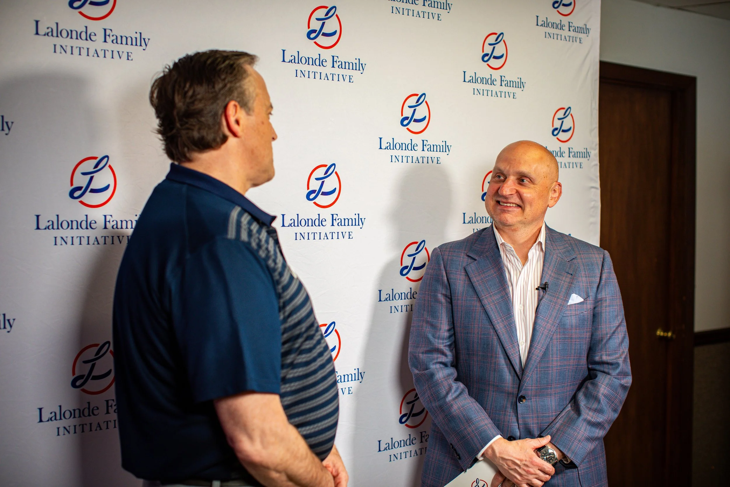 Two men engaged in conversation in front of a backdrop with a logo reading "Lalonde Family Initiative." The man on the left, facing away from the camera, wears a dark blue shirt. The man on the right, facing the camera, is bald, smiling, and dressed in a checkered blazer with a white shirt.