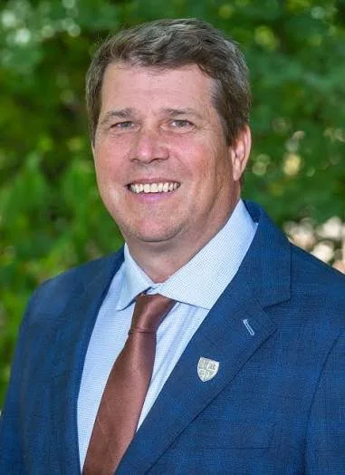 Portrait of a smiling man in a blue suit and brown tie, outside with green foliage in the background.