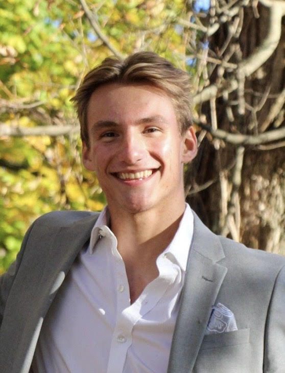A young man in a light gray suit and white shirt smiling outdoors with trees in the background.
