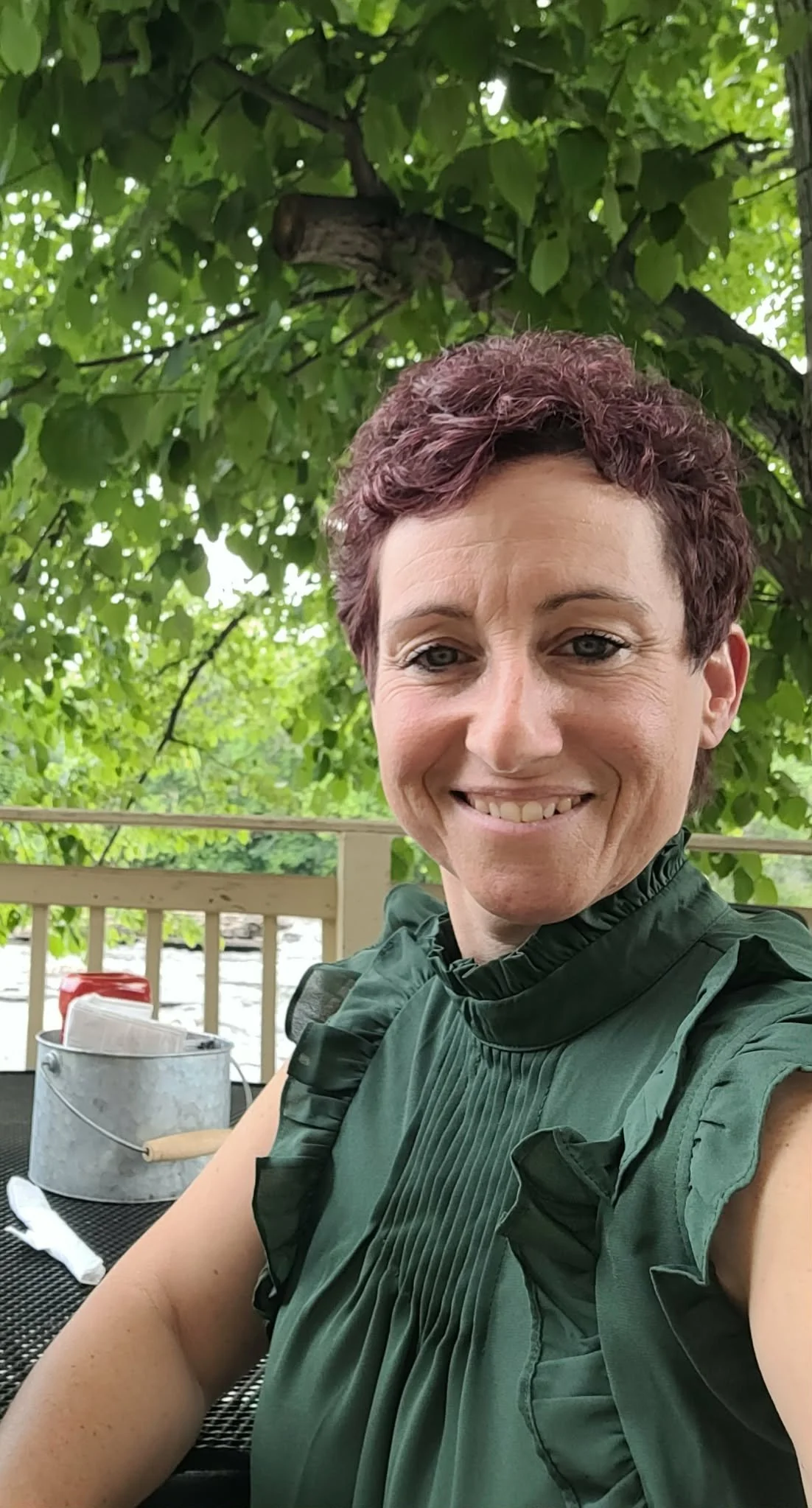 A smiling woman with short, curly red hair taking a selfie outdoors under a leafy tree. She is wearing a dark green sleeveless top with ruffled shoulders, seated at a black outdoor table with a metal bucket and some condiments in the background.