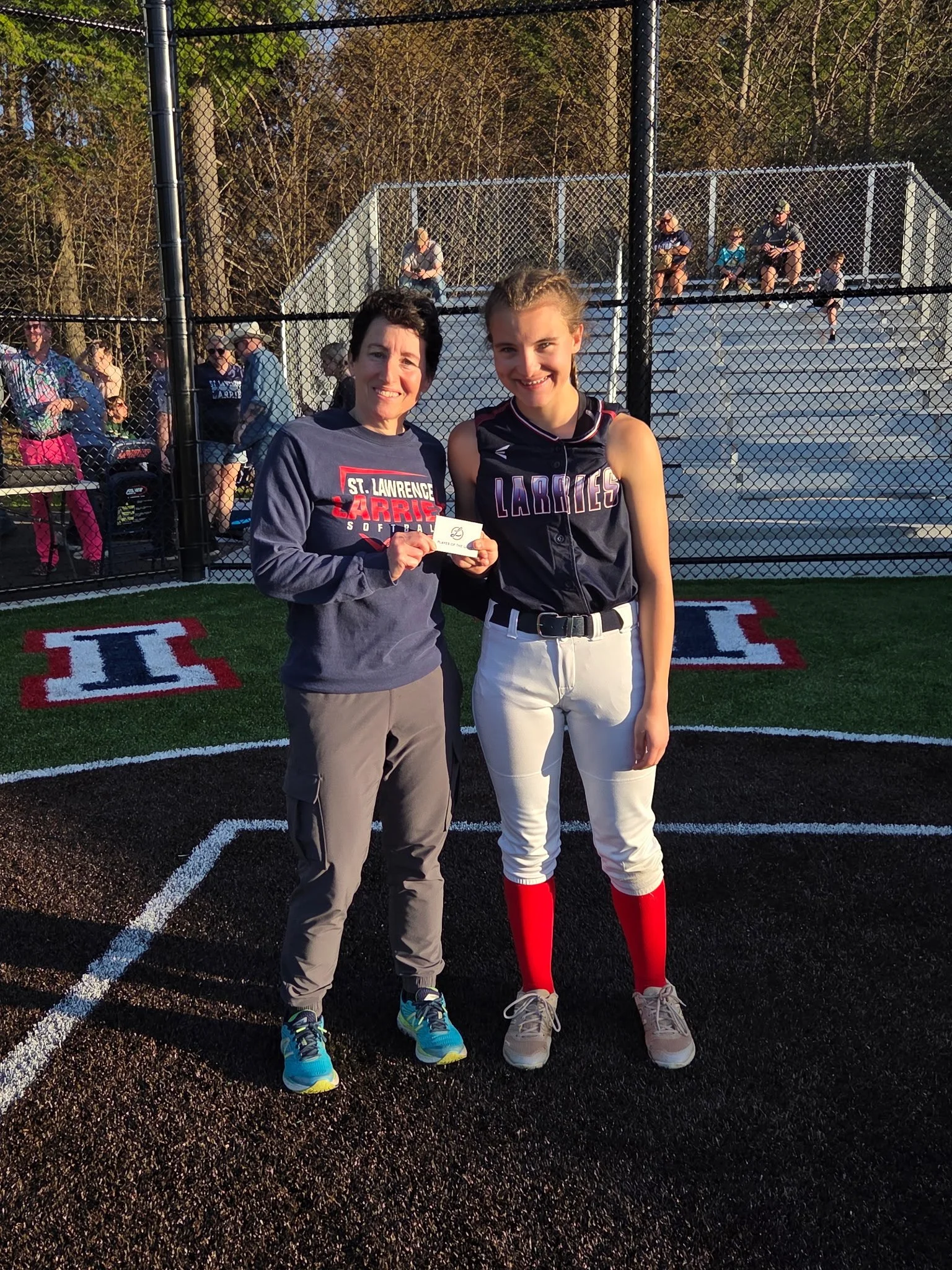 A woman and a girl standing on a baseball field in front of a chain-link fence, with bleachers and other people in the background. The woman is holding a small card, and both are smiling at the camera. The girl is wearing a baseball uniform with red socks, and the woman is dressed casually.
